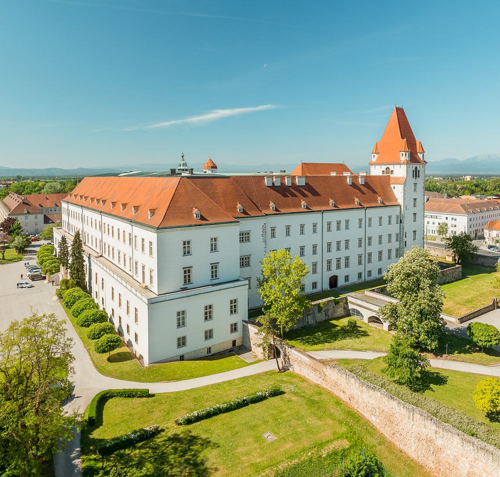 Blick auf eine historische Militärakademie mit rotem Dach und Turm, umgeben von grünen Bäumen und Rasenflächen, im Hintergrund Berge.