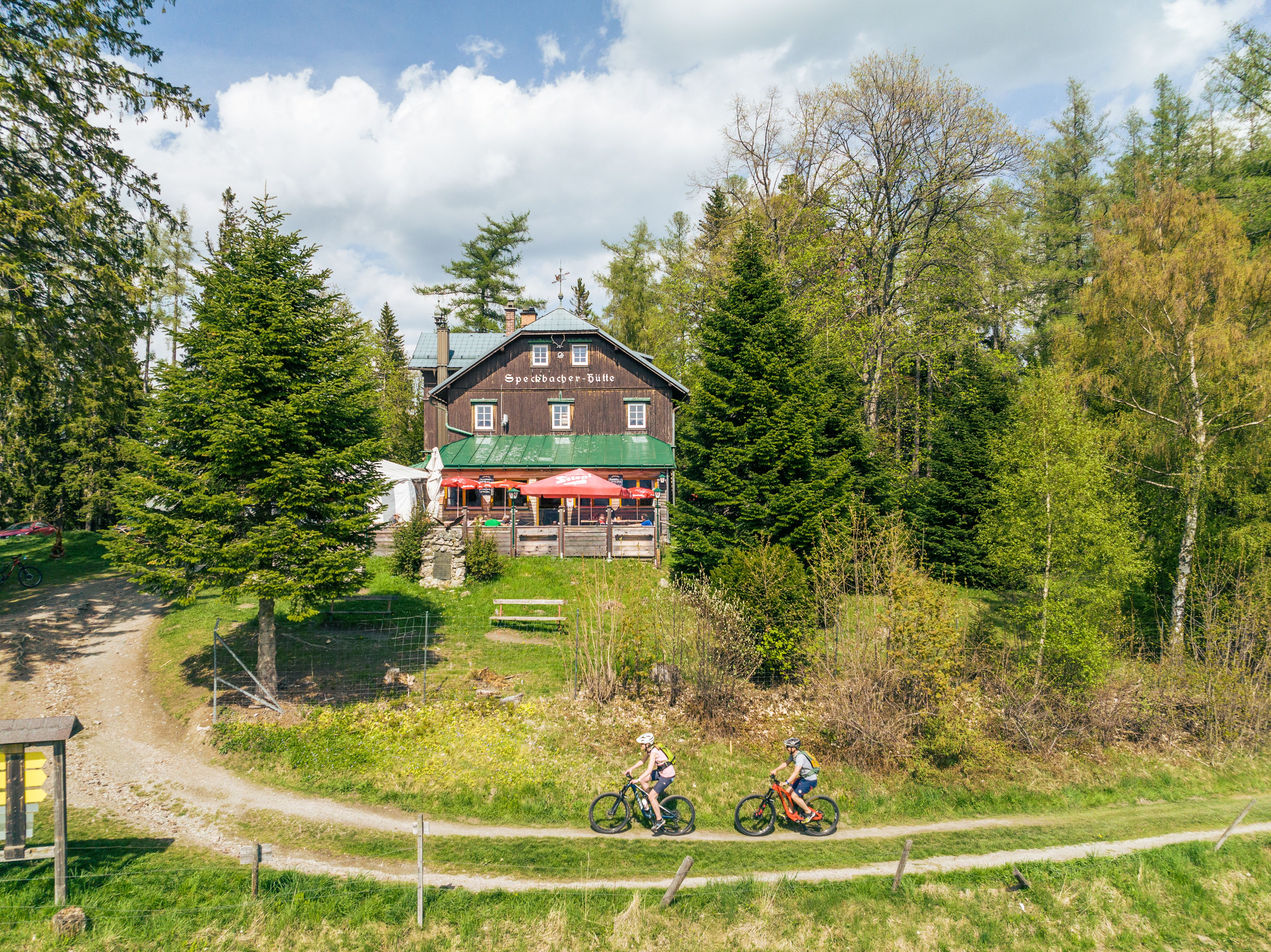 Holzhütte mit der Aufschrift "Speckbacherhütte", rundherum Bäume und zwei Mountainbikefahrer davor