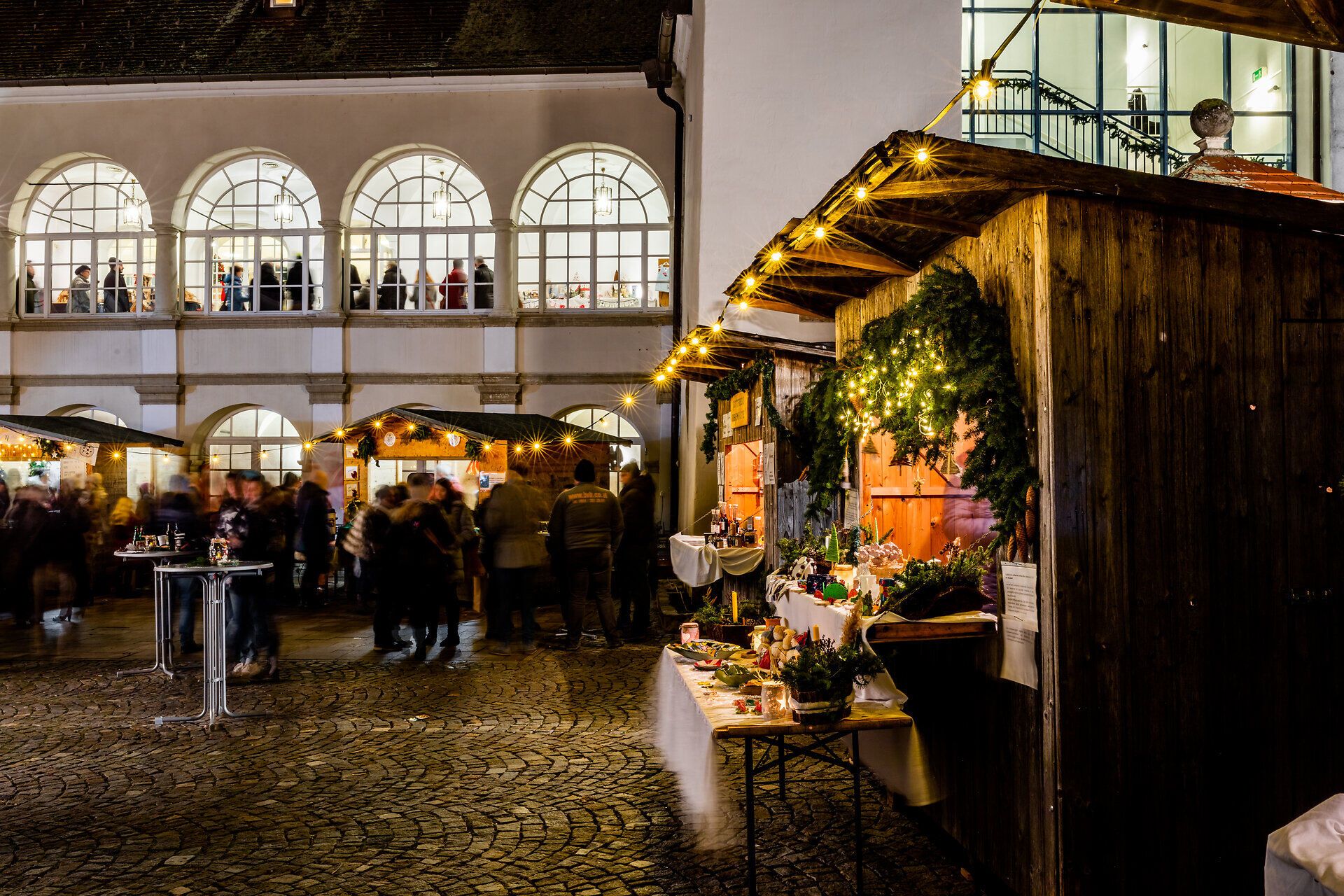 Belebter Innenhof mit Marktständen und warm beleuchteten Fenstern am Abend.