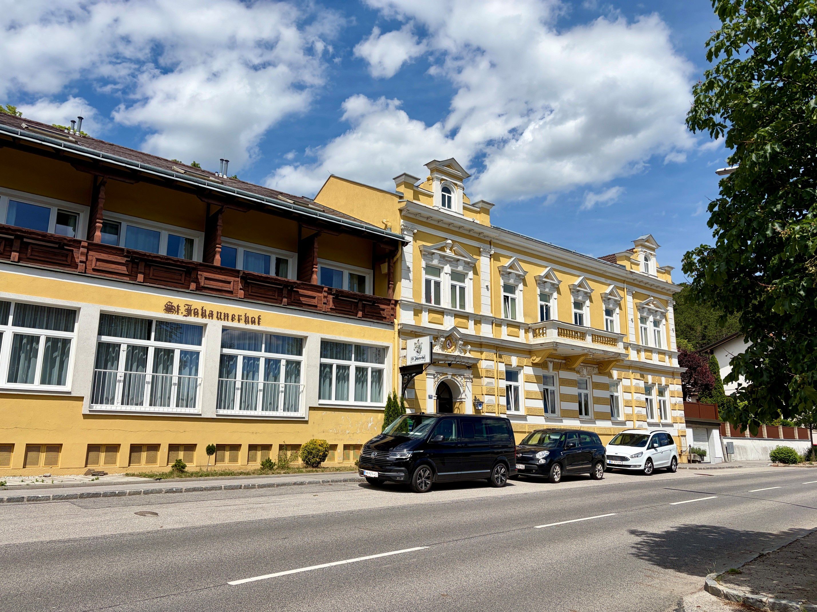 Yellow building with the inscription 'St. Johannerhof' on a street, surrounded by parked cars and trees.