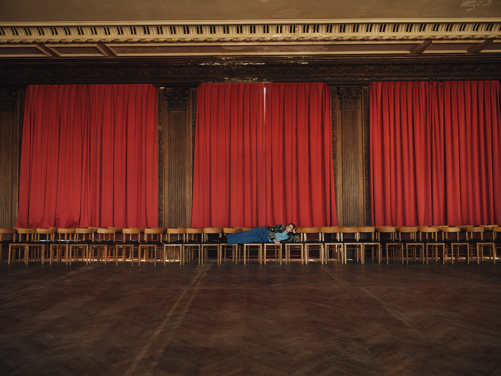 In einem weitläufigen Raum mit roten Vorhängen und einer langen Reihe von Stühlen entsteht eine ruhige, fast melancholische Atmosphäre. Ein einzelner Mensch ruht entspannt auf den Stühlen und genießt die Stille, während das Licht sanft durch die Fenster strömt und die Schönheit des Moments unterstreicht.