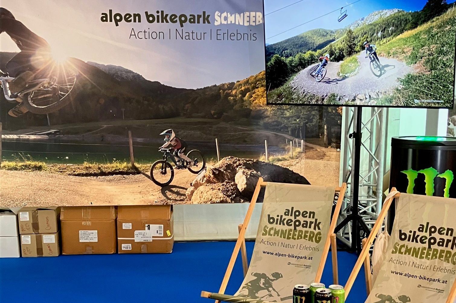 Der Messestand vom Alpen Bikepark Schneeberg bestehend aus großer Fotorückwand, Bildschirm, 2 bedruckten Liegestühlen und einem Tischchen mit Getränken und Prospekten. 