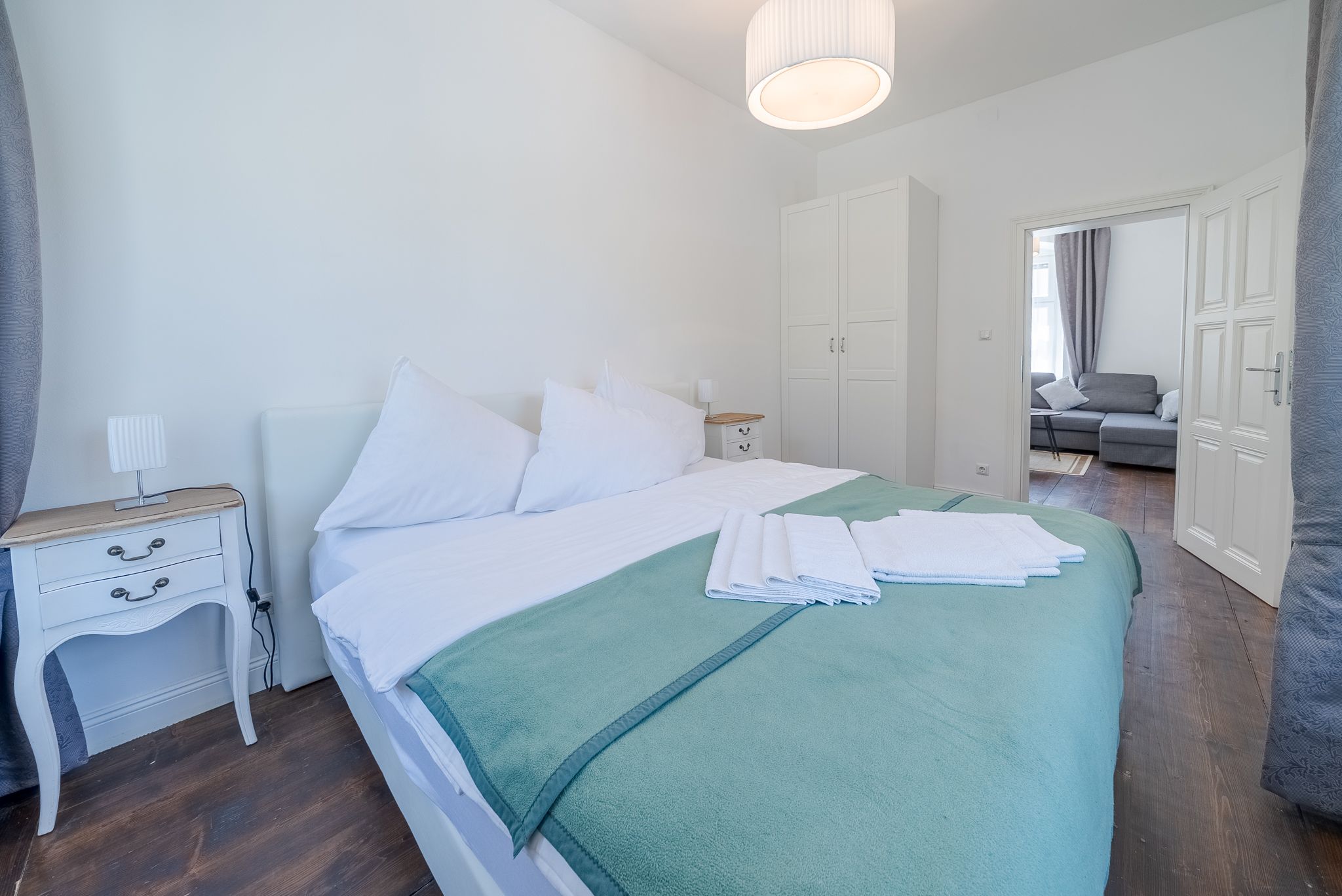 Ein helles Schlafzimmer mit einem großen Bett, weißen Möbeln und einem offenen Durchgang zu einem Wohnzimmer mit grauem Sofa.