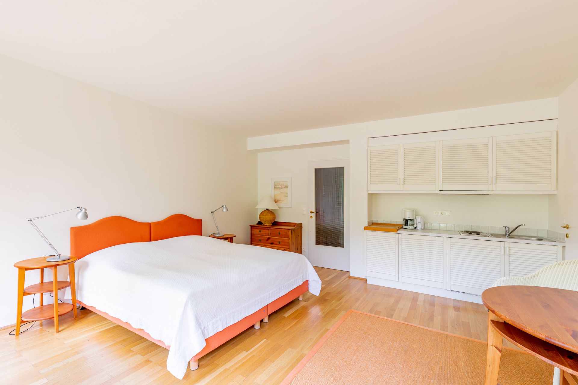 Helles Schlafzimmer mit Doppelbett, Holzmöbeln und kleiner Küchenzeile.