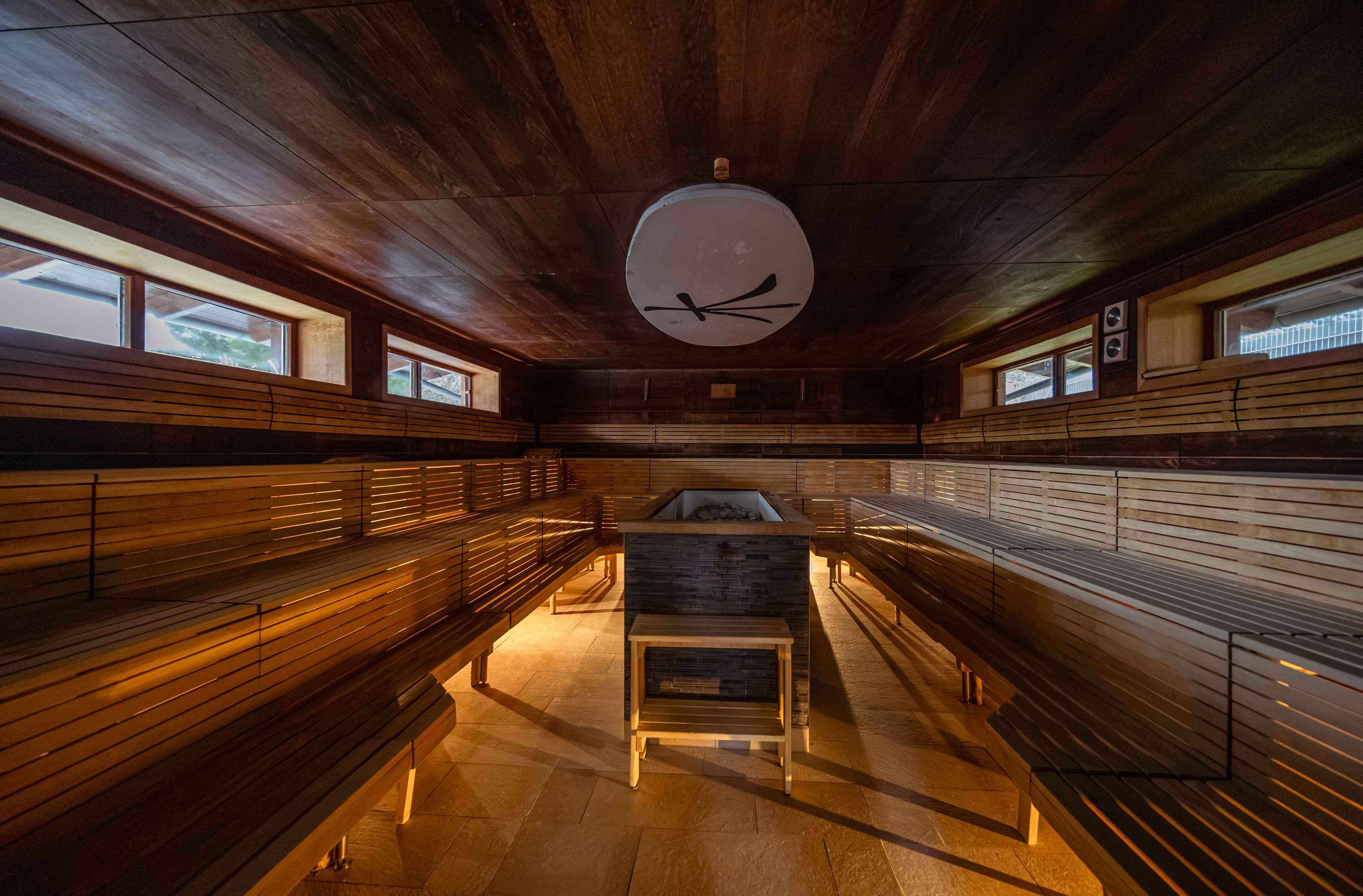 Innenansicht einer Sauna mit Holzbänken und zentralem Ofen.
