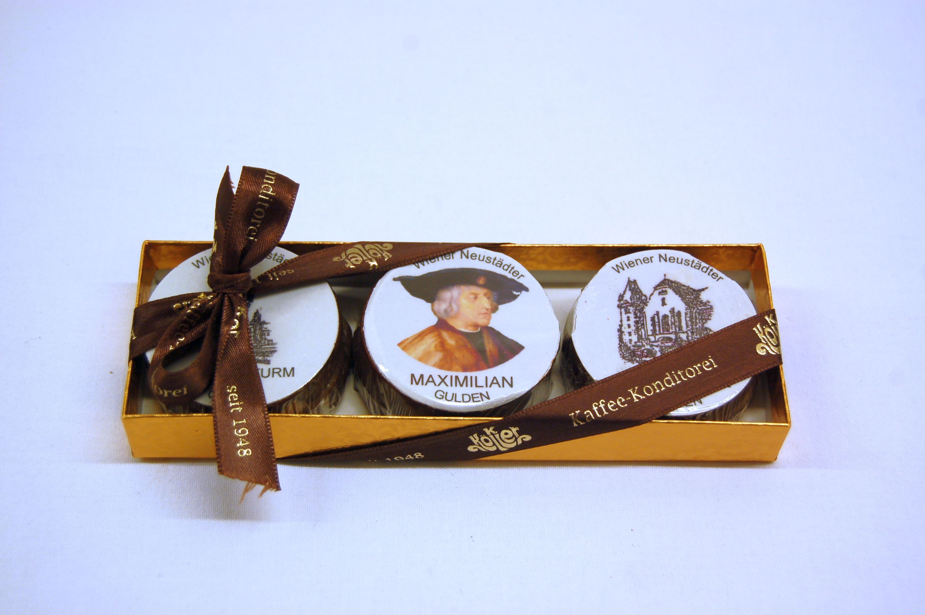Eine goldene Schachtel mit drei Pralinen, verziert mit einem Bild von Maximilian und Wiener Neustadt Motiven, gebunden mit einem braunen Band.