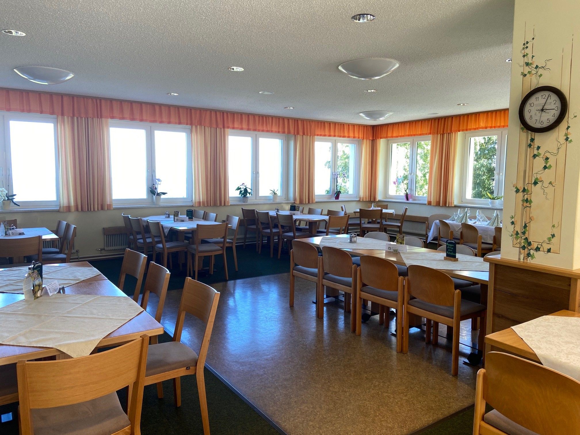 Ein heller Speisesaal mit Holzmöbeln, Tischen und Stühlen, dekoriert mit Pflanzen und einer Wanduhr.