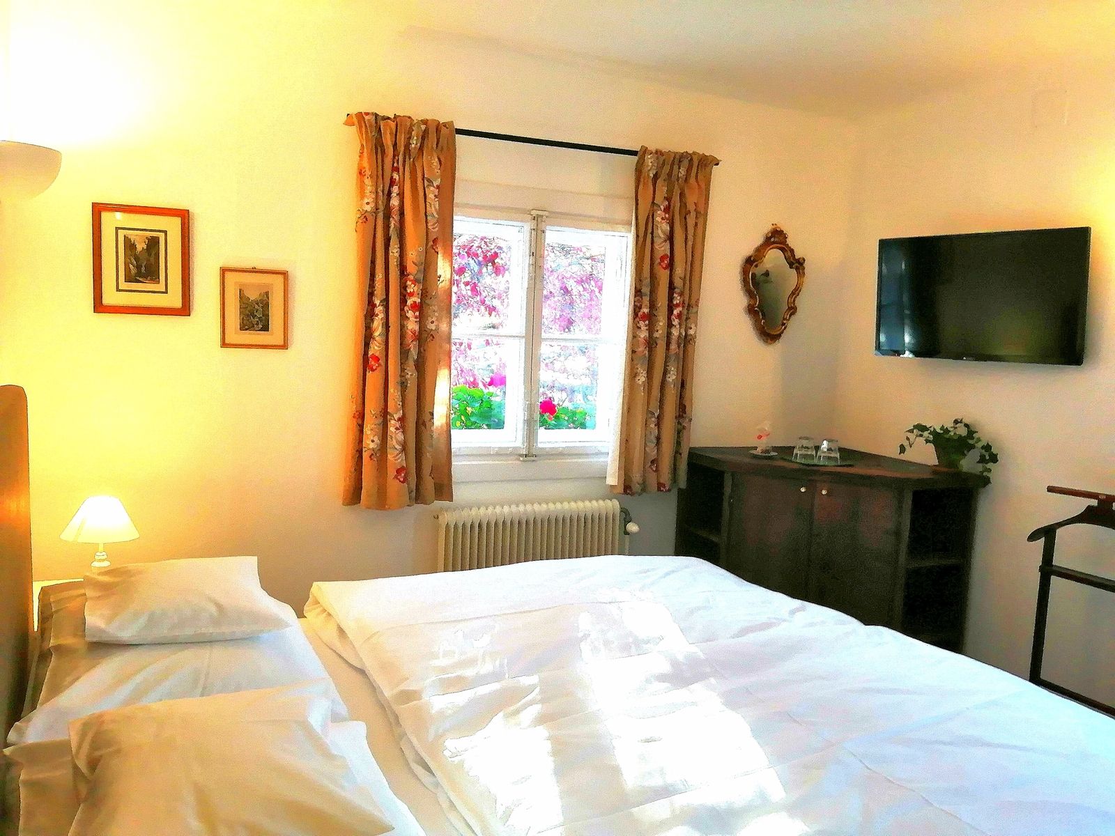 Gemütliches Doppelzimmer mit Bett, TV, Fenster mit Vorhängen, Bildern an der Wand und einem kleinen Schrank.