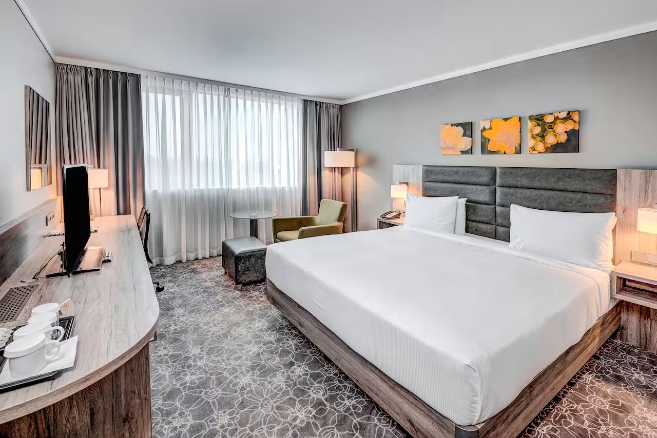 Großes Zimmer mit Doppelbett, Sitzmöglichkeit, Fernseher, grauen Vorhängen und gemustertem Teppich.