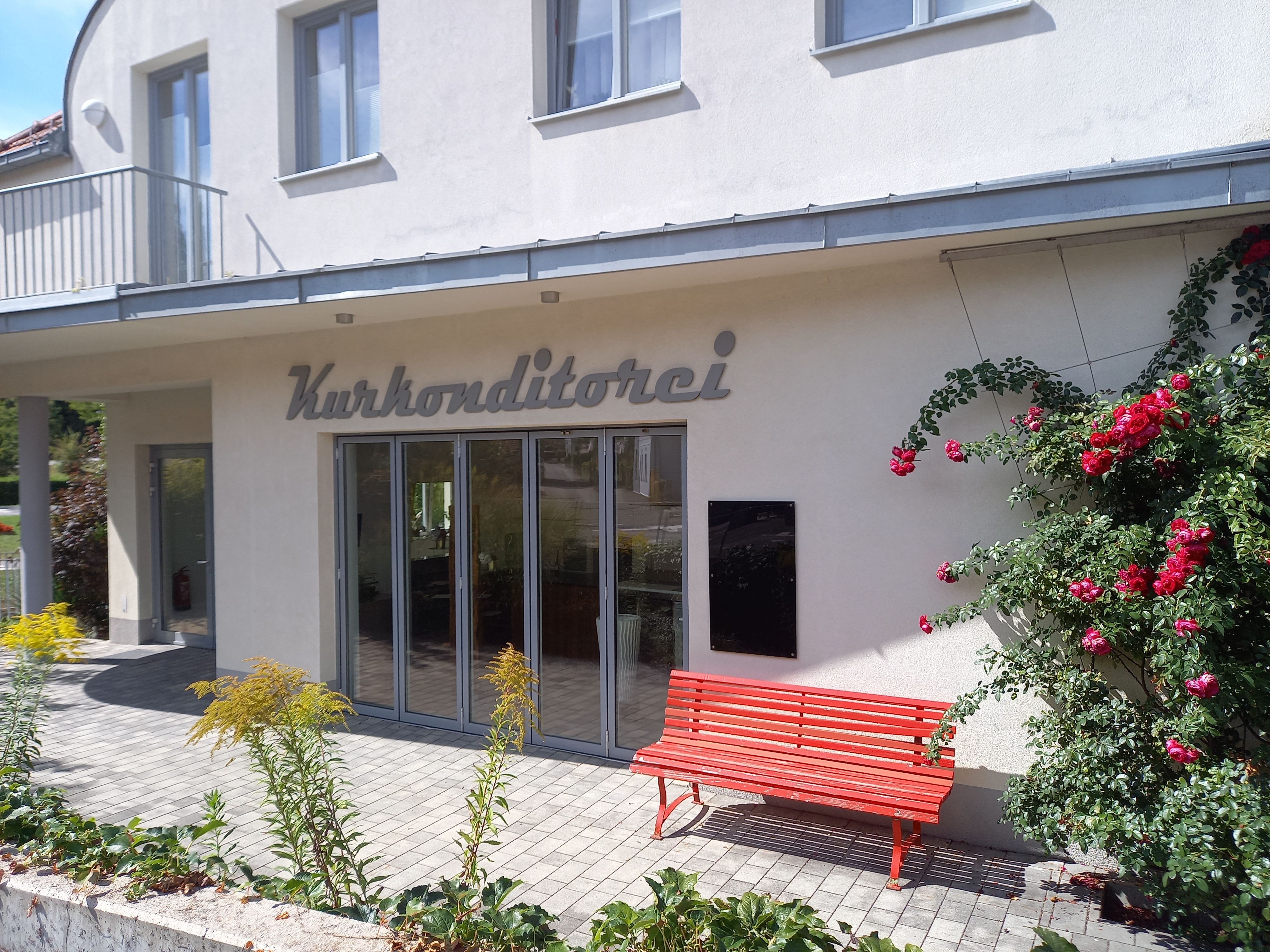 Außenansicht eines Gebäudes mit der Aufschrift 'Kurkonditorei', roter Bank und blühenden Rosen.