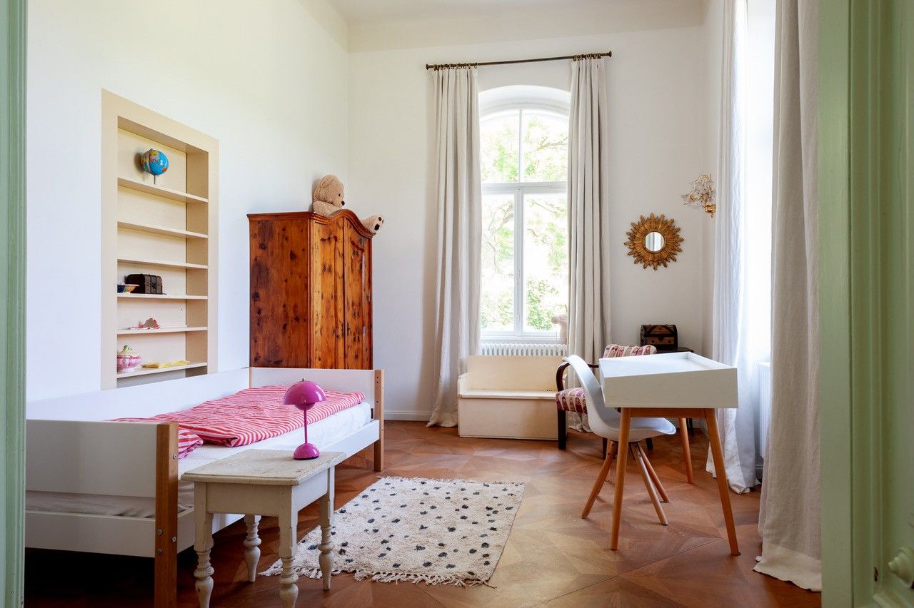 Ein helles Kinderzimmer mit Holzboden, einem Bett, einem Schreibtisch und einem Schrank. Ein Fenster mit Vorhängen lässt Tageslicht herein.