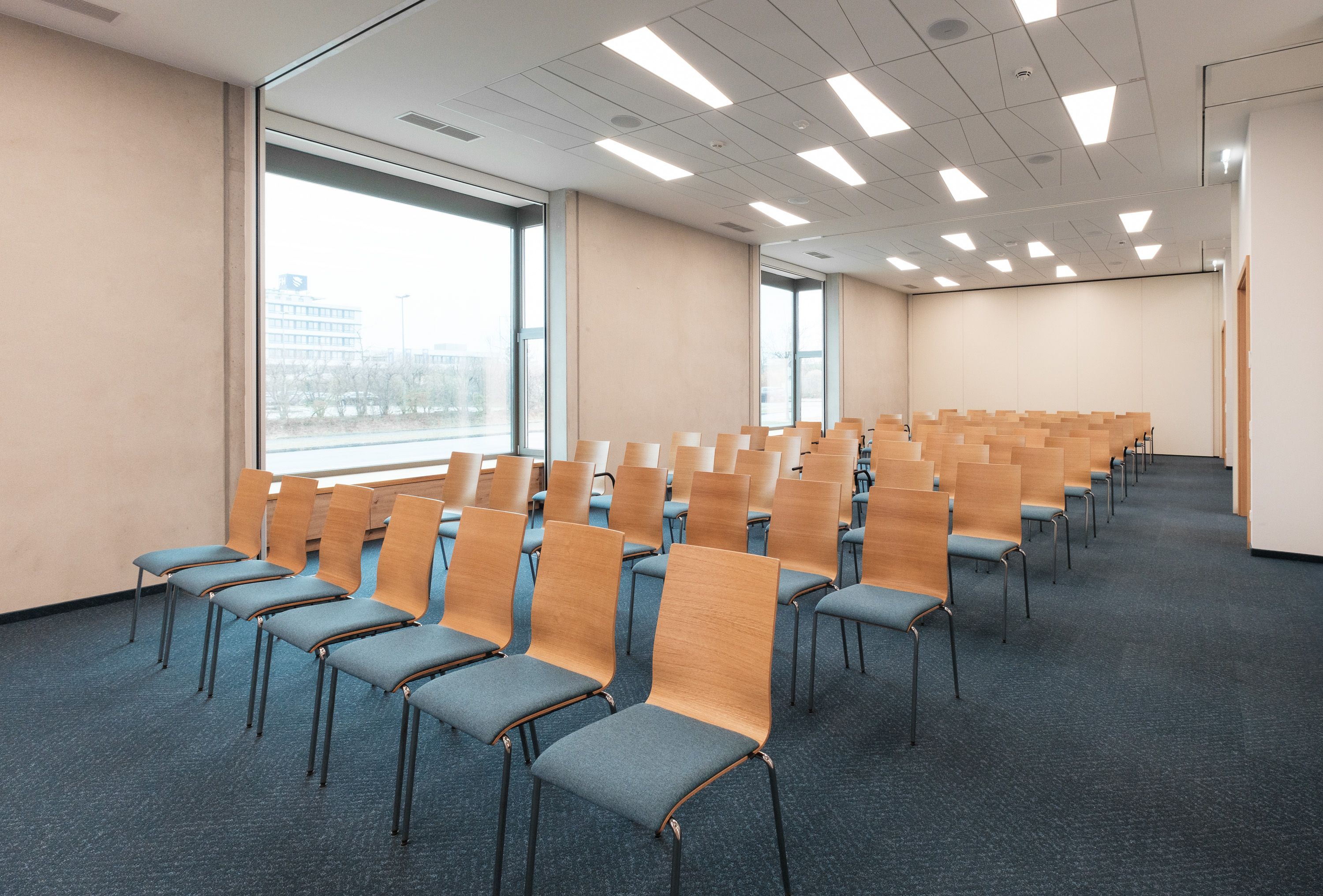 Theaterbestuhlung im Seminarraum. Blauer Teppichoden, Stühle mit Sitzkissen und viel Tageslicht.