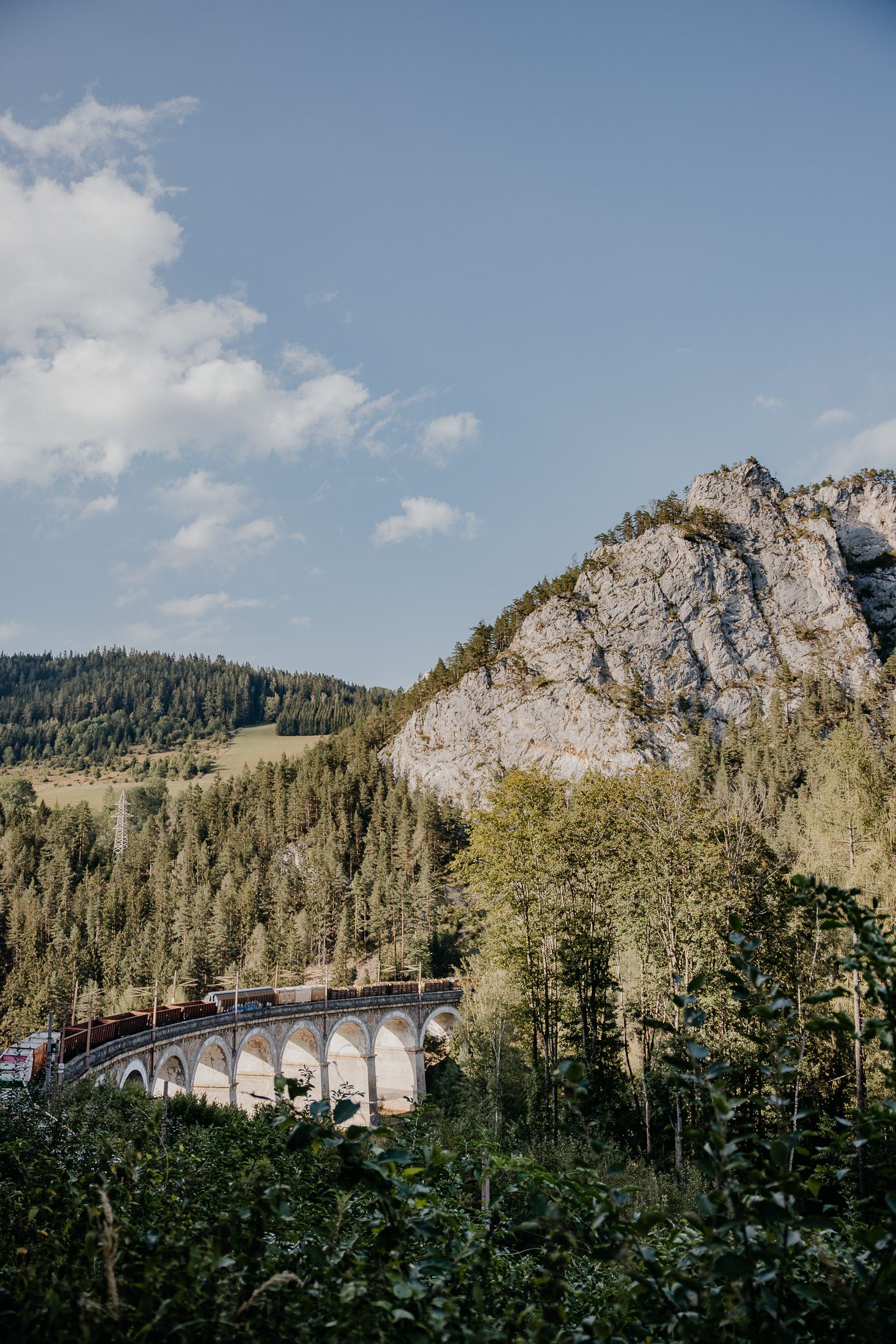 Semmeringbahnwanderweg mit Steinviadukt der Semmeringbahn, bewaldeten Hängen und Felsformation unter blauem Himmel.
