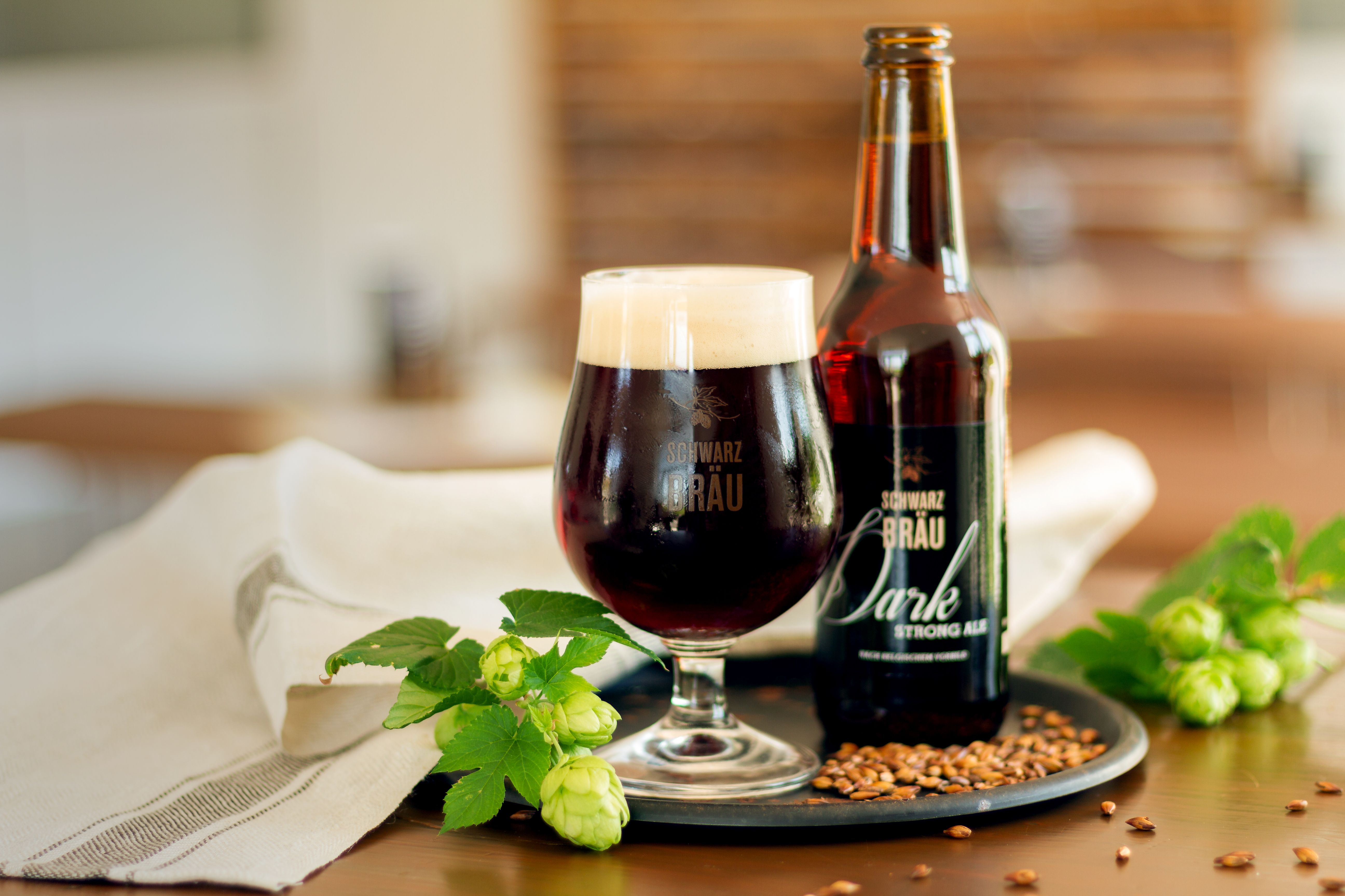 Eine Flasche und ein Glas mit Dark Strong Ale von Schwarzbräu auf einem Tablett, daneben Hopfen.