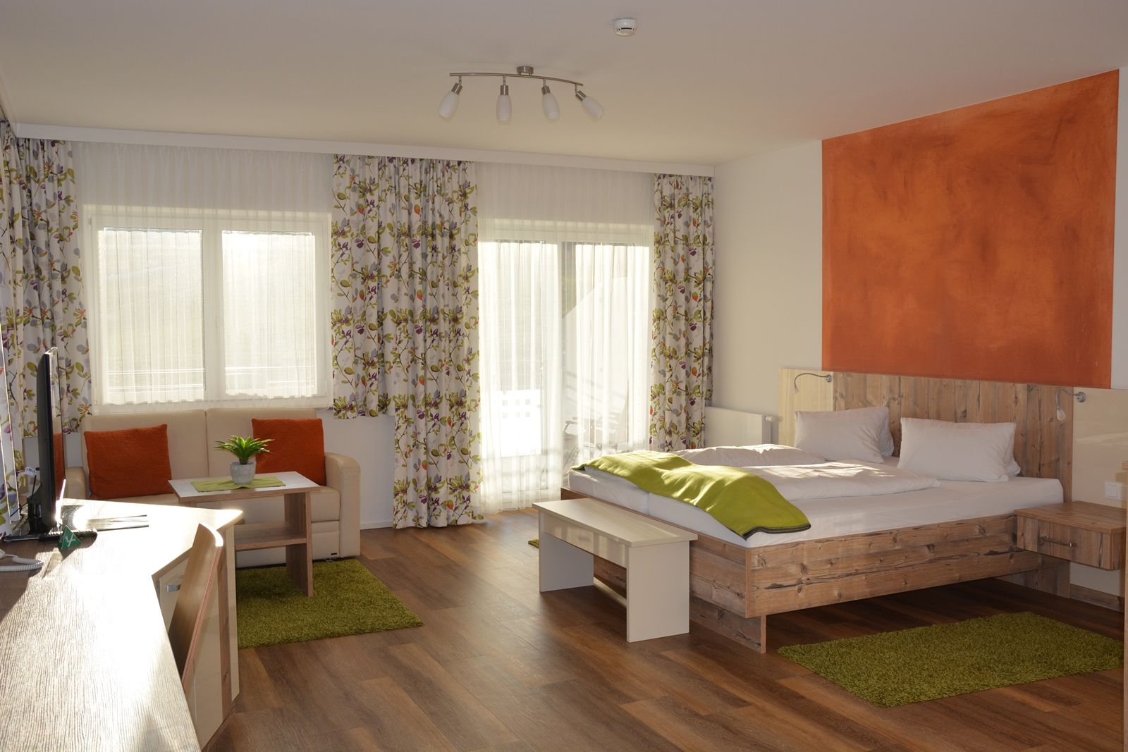 Ein modernes Doppelzimmer mit Holzboden, einem Doppelbett, Sofa, Tisch und großen Fenstern mit Vorhängen.