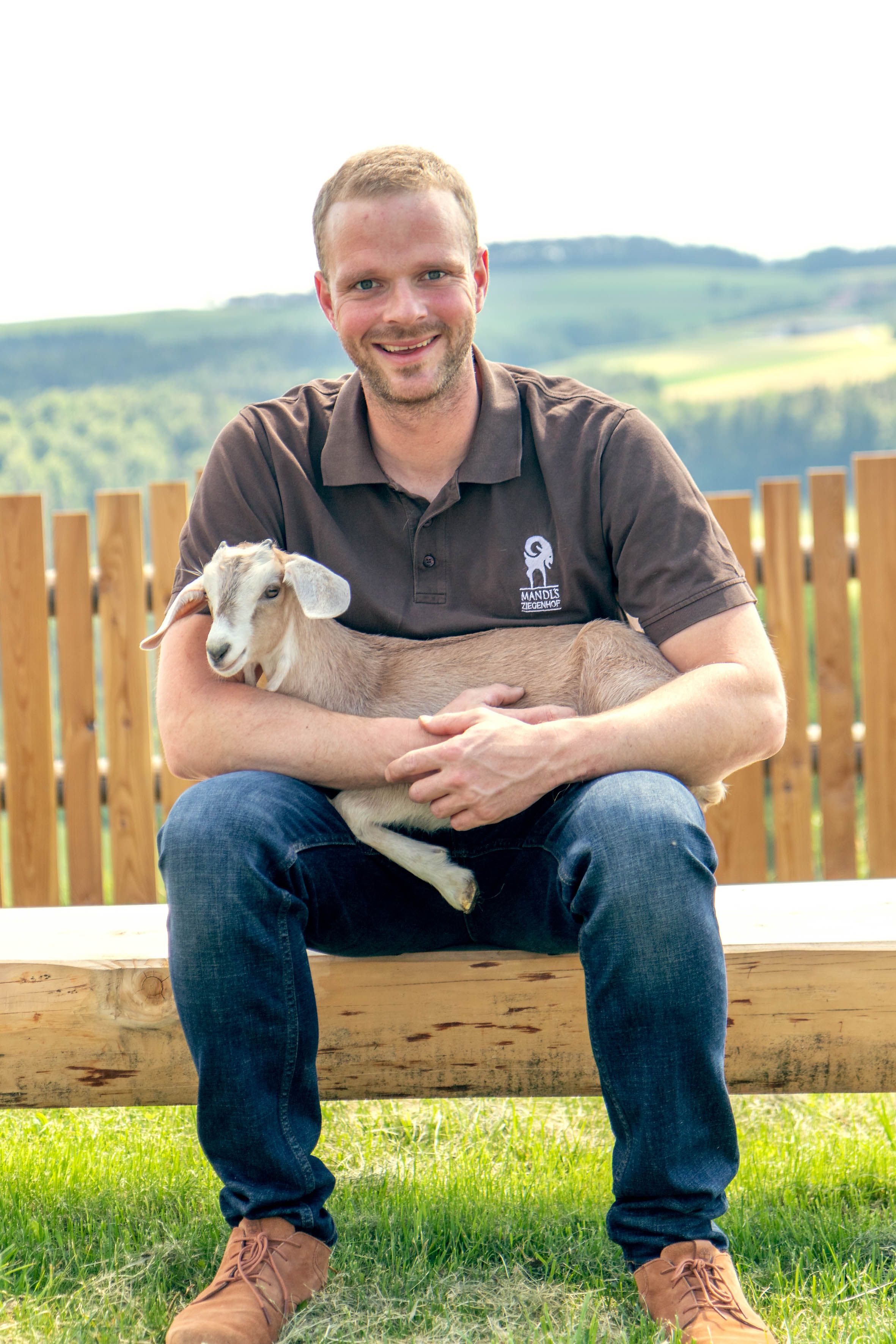 Ein Mann sitzt auf einer Holzbank, hält eine Ziege und lächelt in die Kamera.