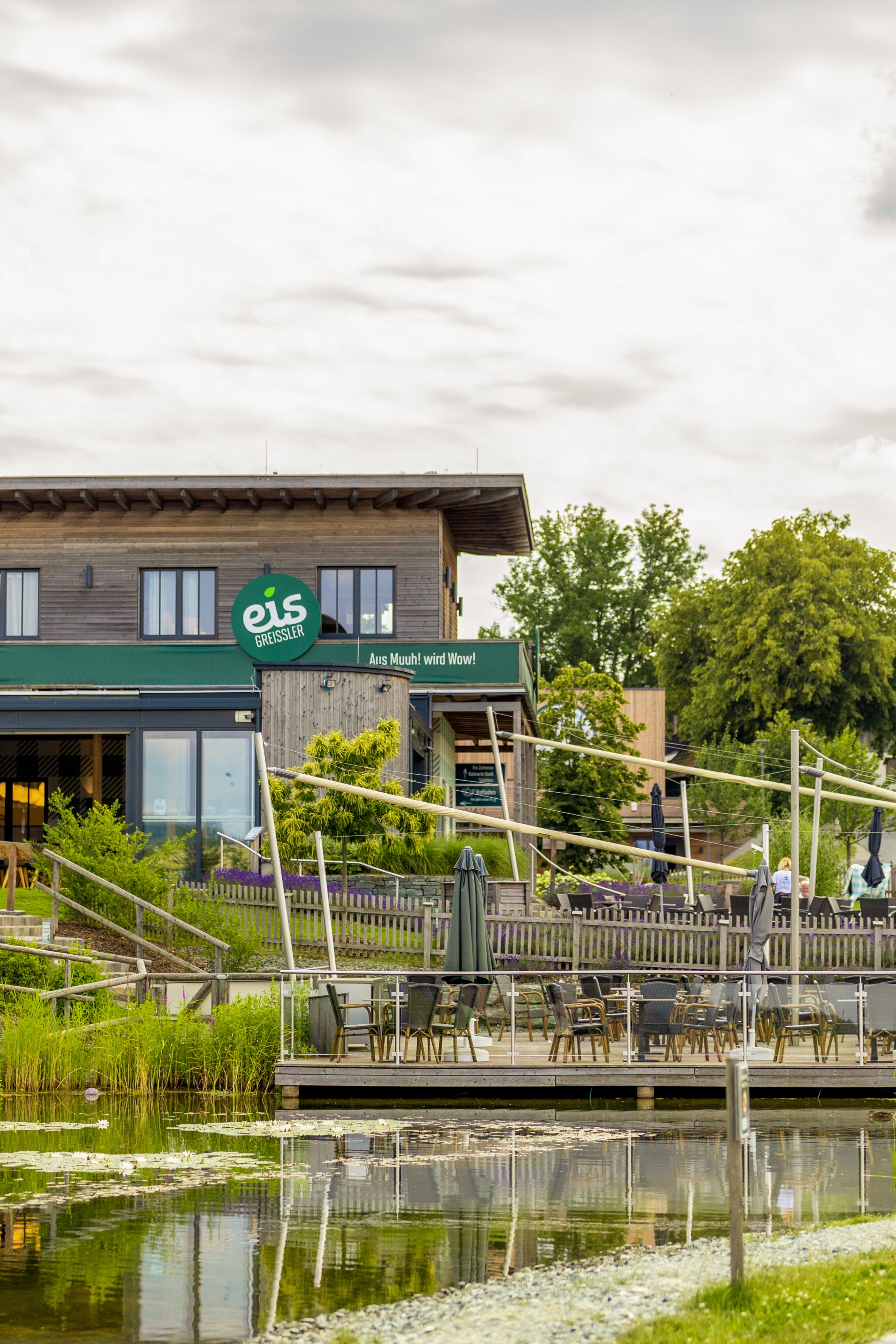 Außenansicht der Eis-Greissler-Welt mit Holzgebäude, See und Terasse mit Tischen, Stühlen und Sonnenschirmen.