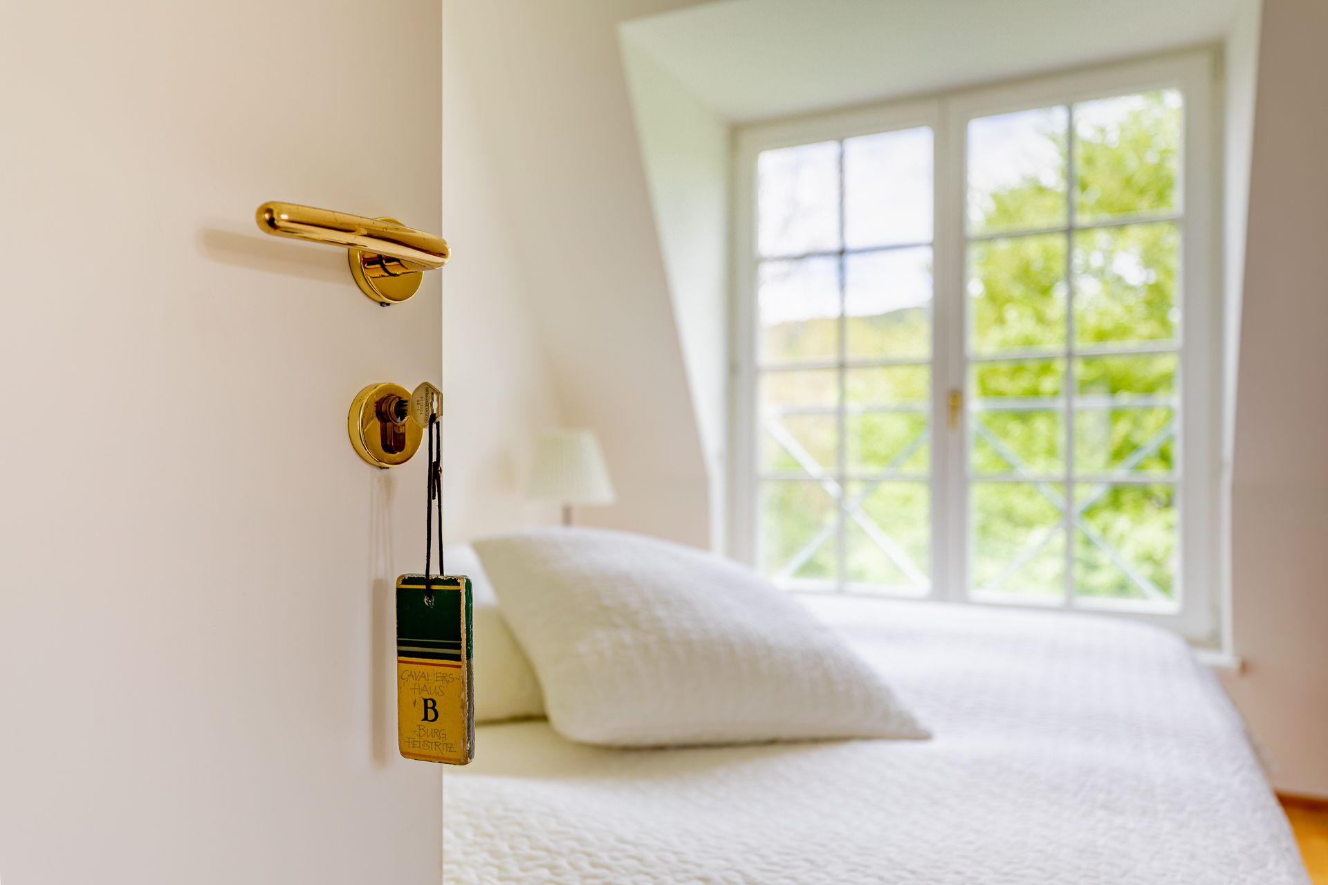 Offene Tür mit Schlüssel und Schild, dahinter ein Bett und Fenster.