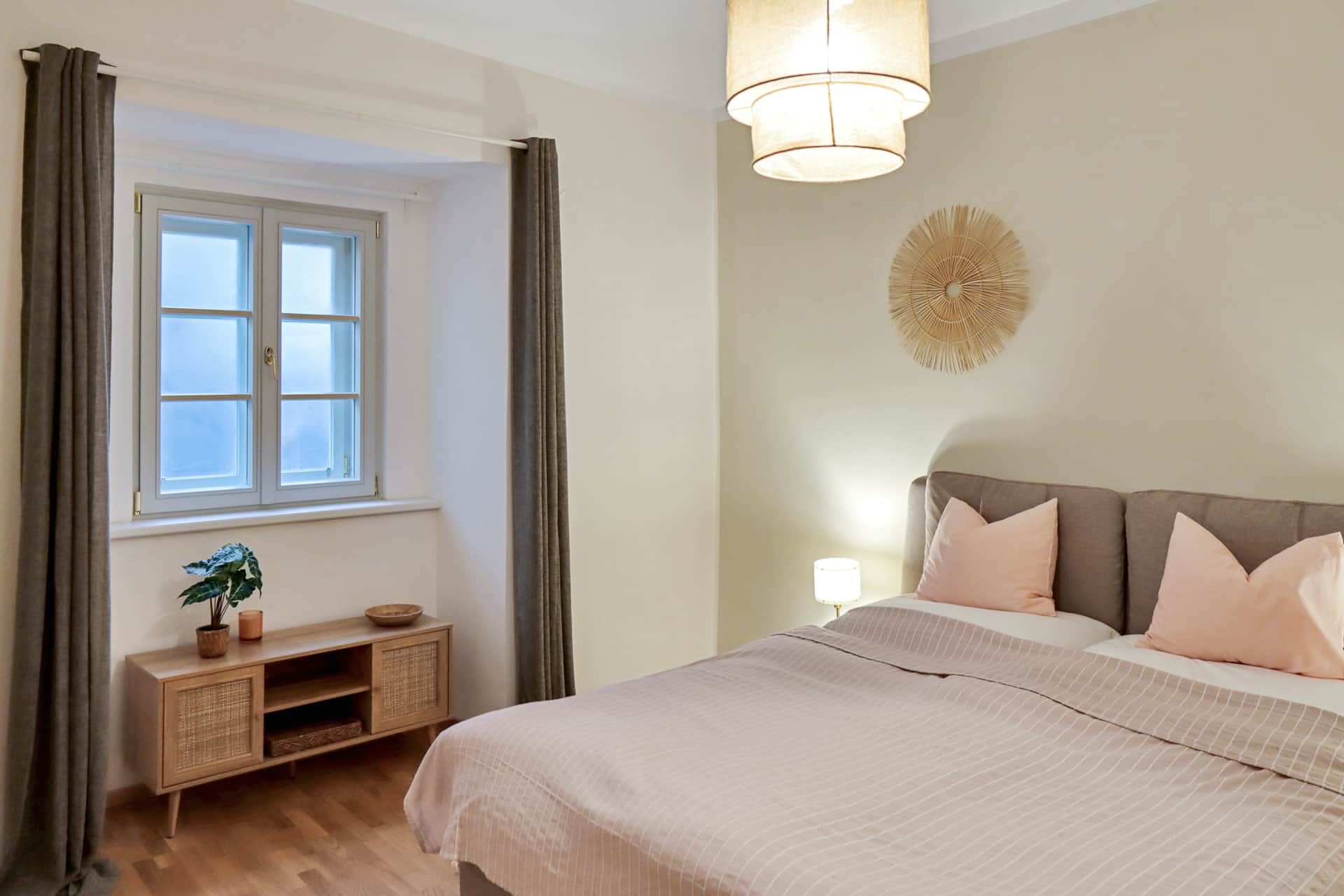 Schlafzimmer mit Doppelbett, Fenster, braunen Vorhängen, Sideboard und Nachtkästchen mit Lampe.