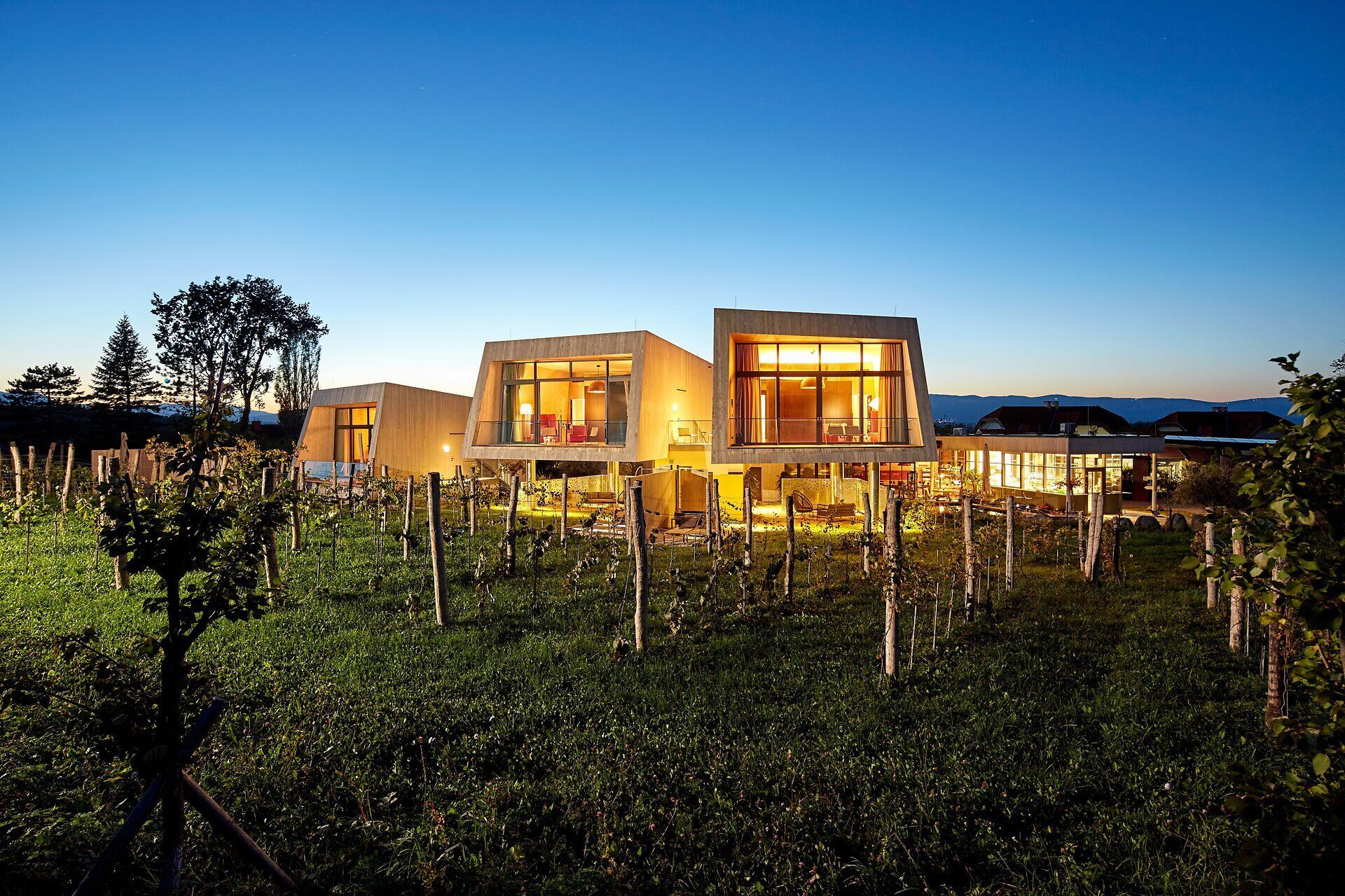 Inmitten der sanften Hügel der Wiener Alpen erstrahlen die modernen Gartenlofts in warmem Licht, während der Abendhimmel in sanften Blau- und Orangetönen leuchtet. Umgeben von üppigen Wiesen und sanften Weinreben, lädt diese idyllische Kulisse zum Entspannen und Genießen ein.