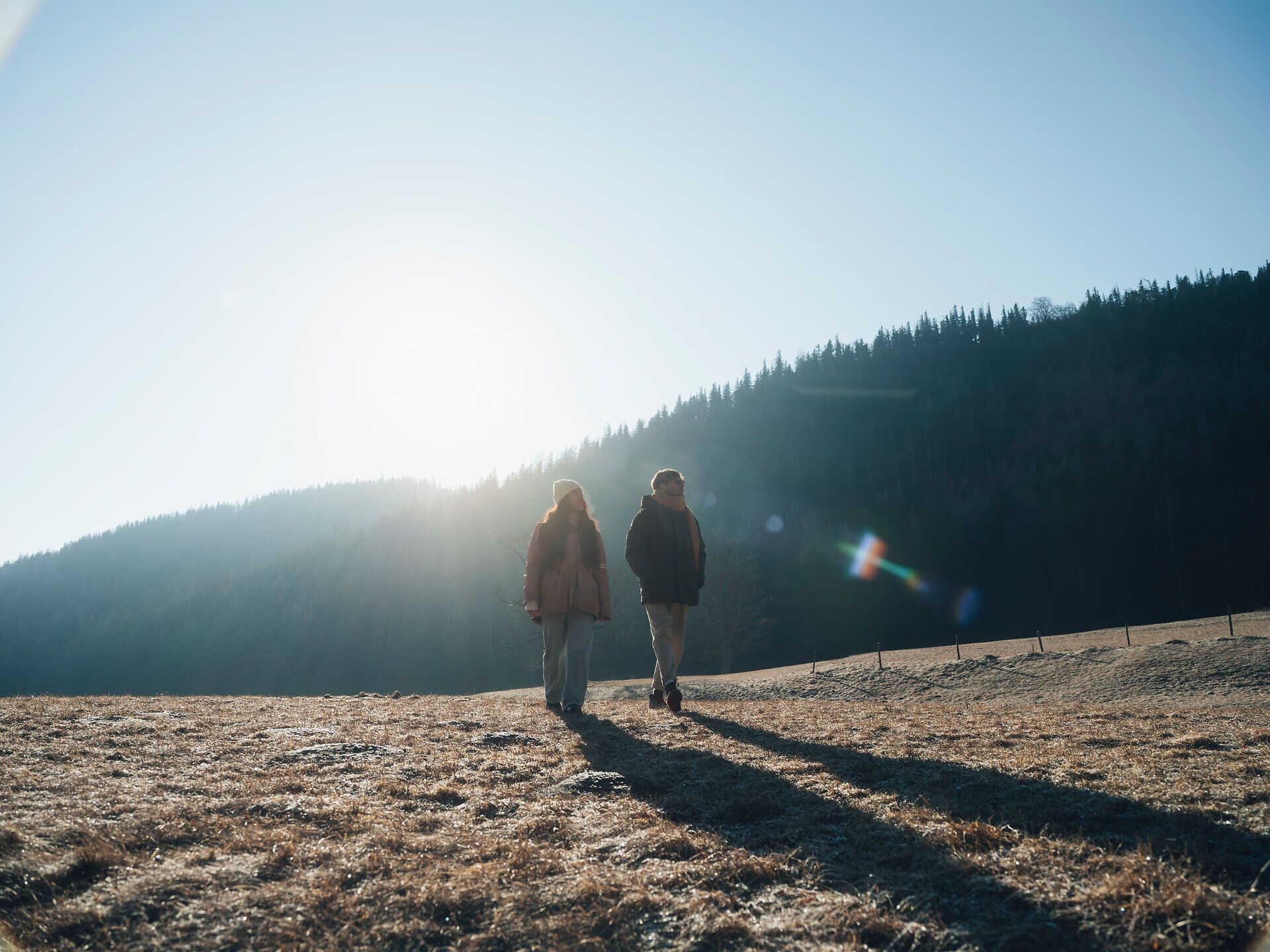 In der winterlichen Landschaft der Wiener Alpen wandern zwei Personen durch die frostige Wiese, während die Sonne sanft über den Horizont strahlt. Die klare, kalte Luft und die schneebedeckten Berge im Hintergrund schaffen eine friedliche und einladende Atmosphäre für Naturliebhaber.