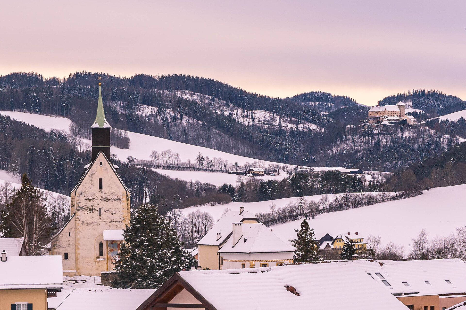 Blick auf die Wehrkirche in Bad Schönau über schneebedeckte Dächer sowie im Hintergrund auf das Schloss Krumbach.