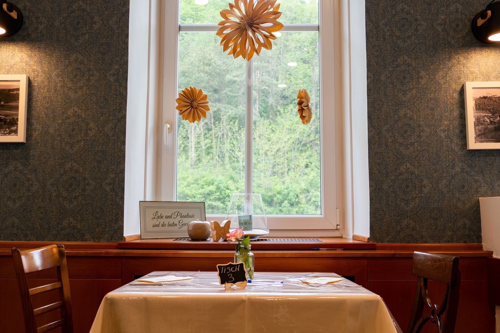 Ein Tisch in einem Restaurant mit Fensterblick, dekoriert mit Papierblumen und einem Schild mit der Aufschrift 'Liebe und Phantasie sind die besten Gewürze'.