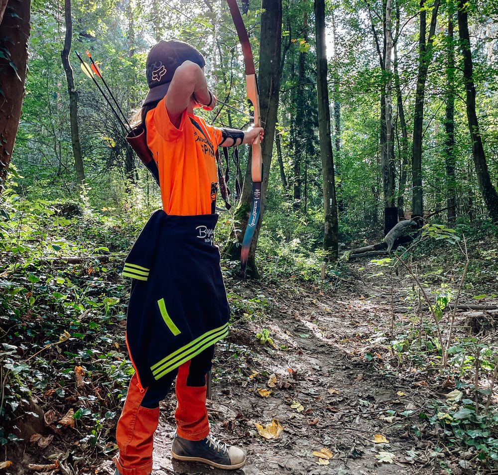 Ein Junge im Wald mit dem Rücken zum Betrachter, er richtet mit guter Körperspannung Pfeil und Bogen auf ein Ziel in der Ferne. Er trägt Leuchtfarben und hat noch 2 Pfeile im Köcher.