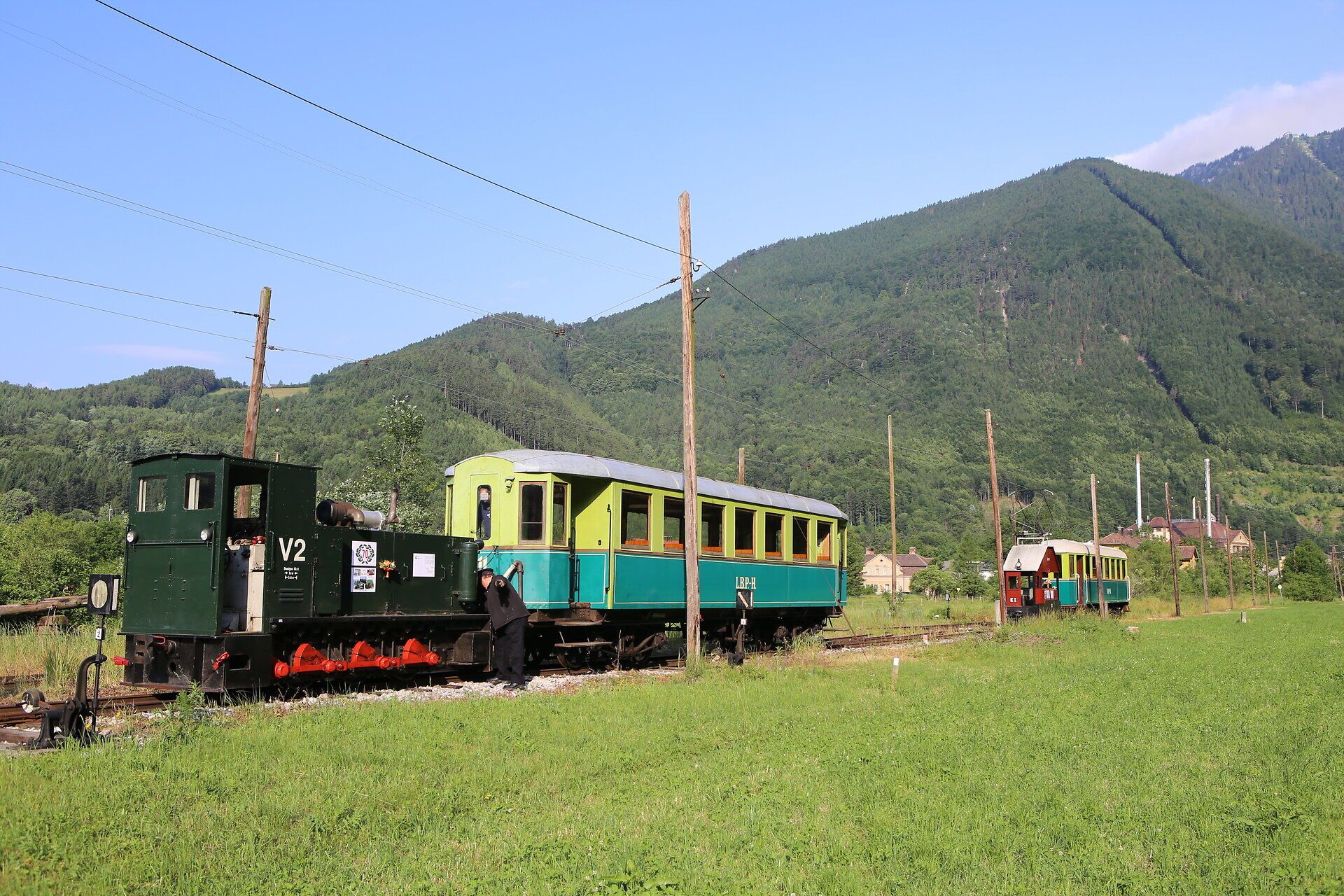 Historische Lokomotive mit Personenwagen steht auf einer Wiese vor einer hügeligen Bergkulisse.