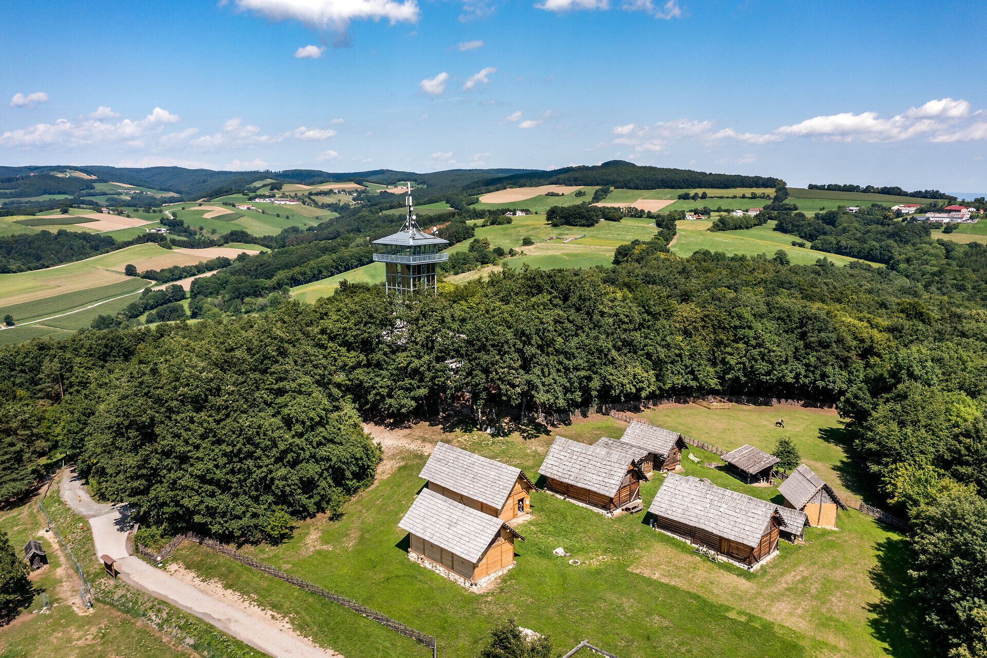 Luftansicht des Keltendorf Schwarzenbach mit den Häusern auf der Wiese und dem Aussichtsturm umgeben von Wald, Feldern und den Hügeln der Buckligen Welt.