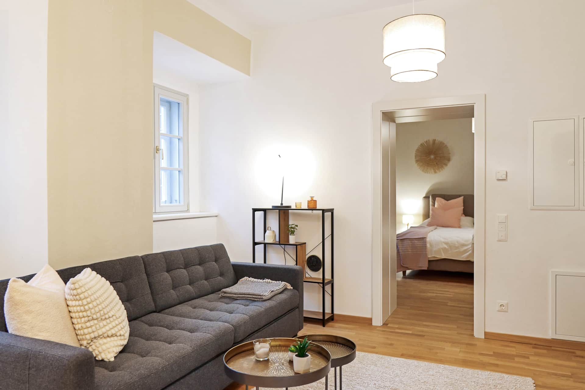 Heller Wohnbereich mit grauer Couch und Wohnzimmertisch mit Blick ins Schlafzimmer