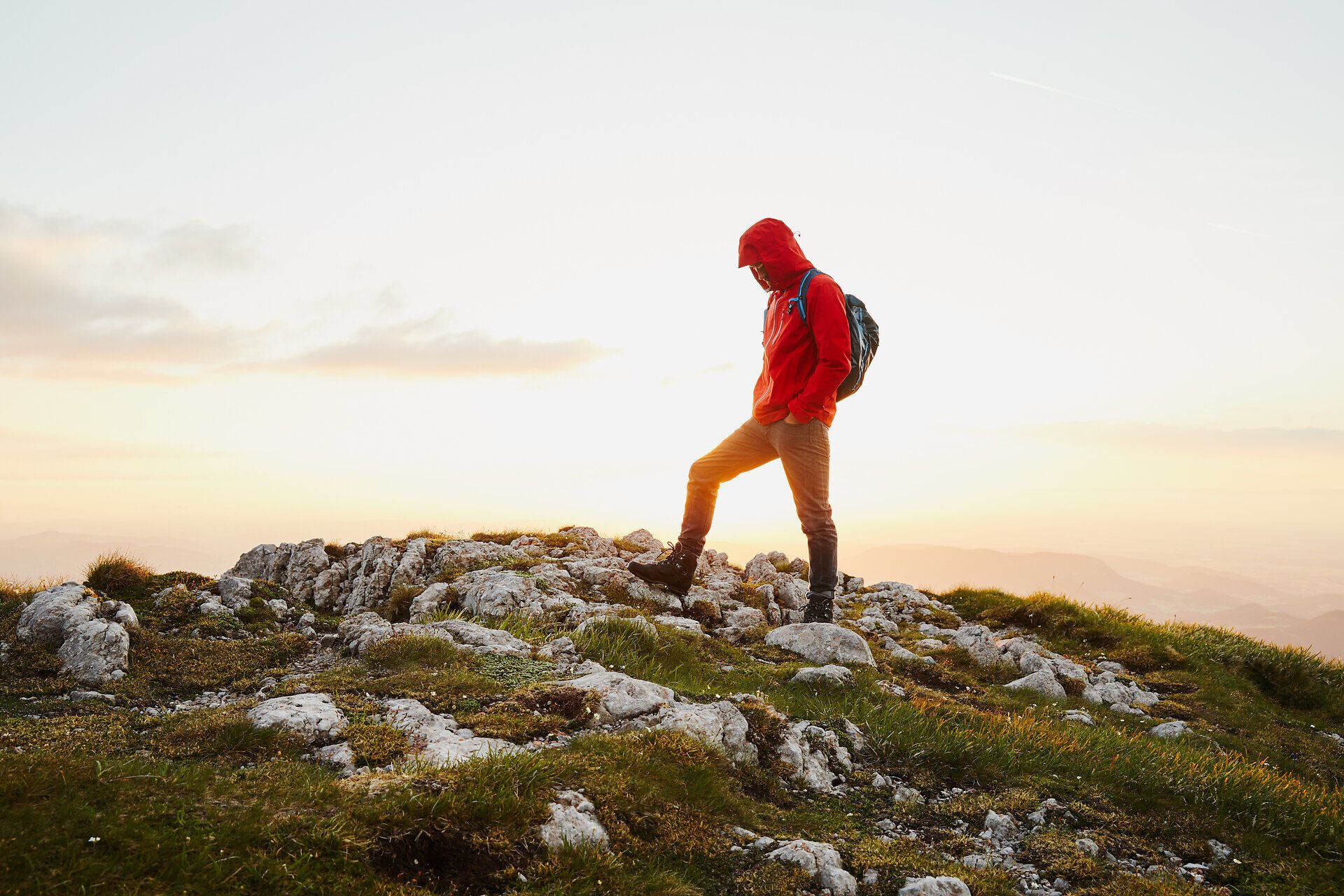 Ein Wanderer steht auf einem felsigen Gipfel und genießt den atemberaubenden Blick auf die umliegenden Berge, während die Sonne langsam am Horizont aufgeht. Die frische Bergluft und die sanften Farben des Morgens schaffen eine friedliche Atmosphäre, die zum Verweilen einlädt.