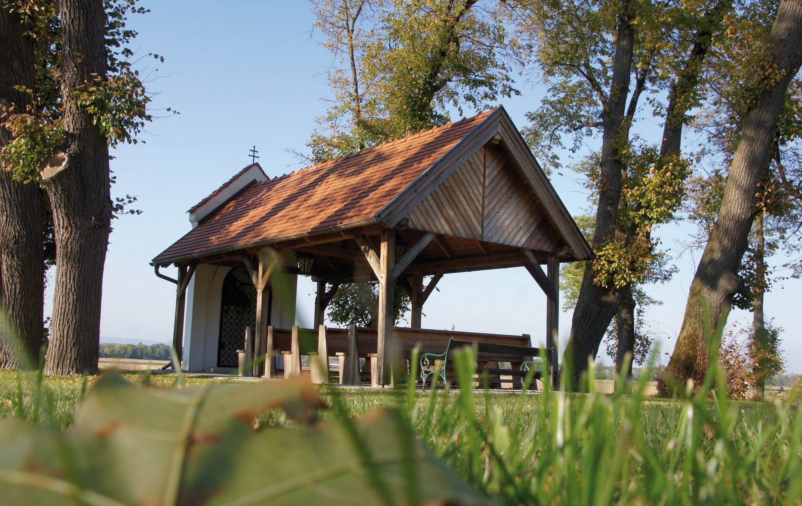Kleine Kapelle mit Holzdach und Bänken, umgeben von Bäumen und Wiese.