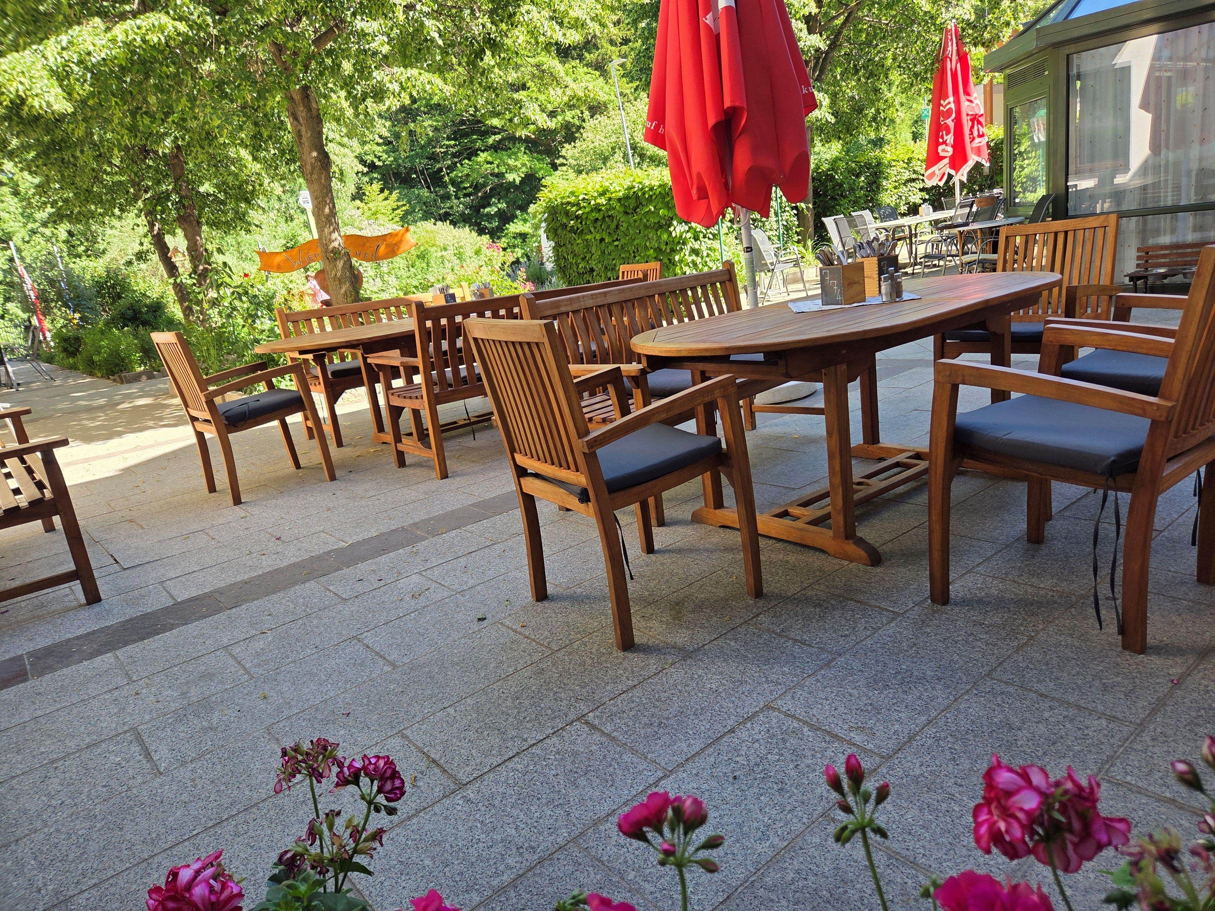 Ein gemütlicher Gastgarten mit Holzmöbeln und roten Sonnenschirmen, umgeben von grünen Bäumen und Blumen.