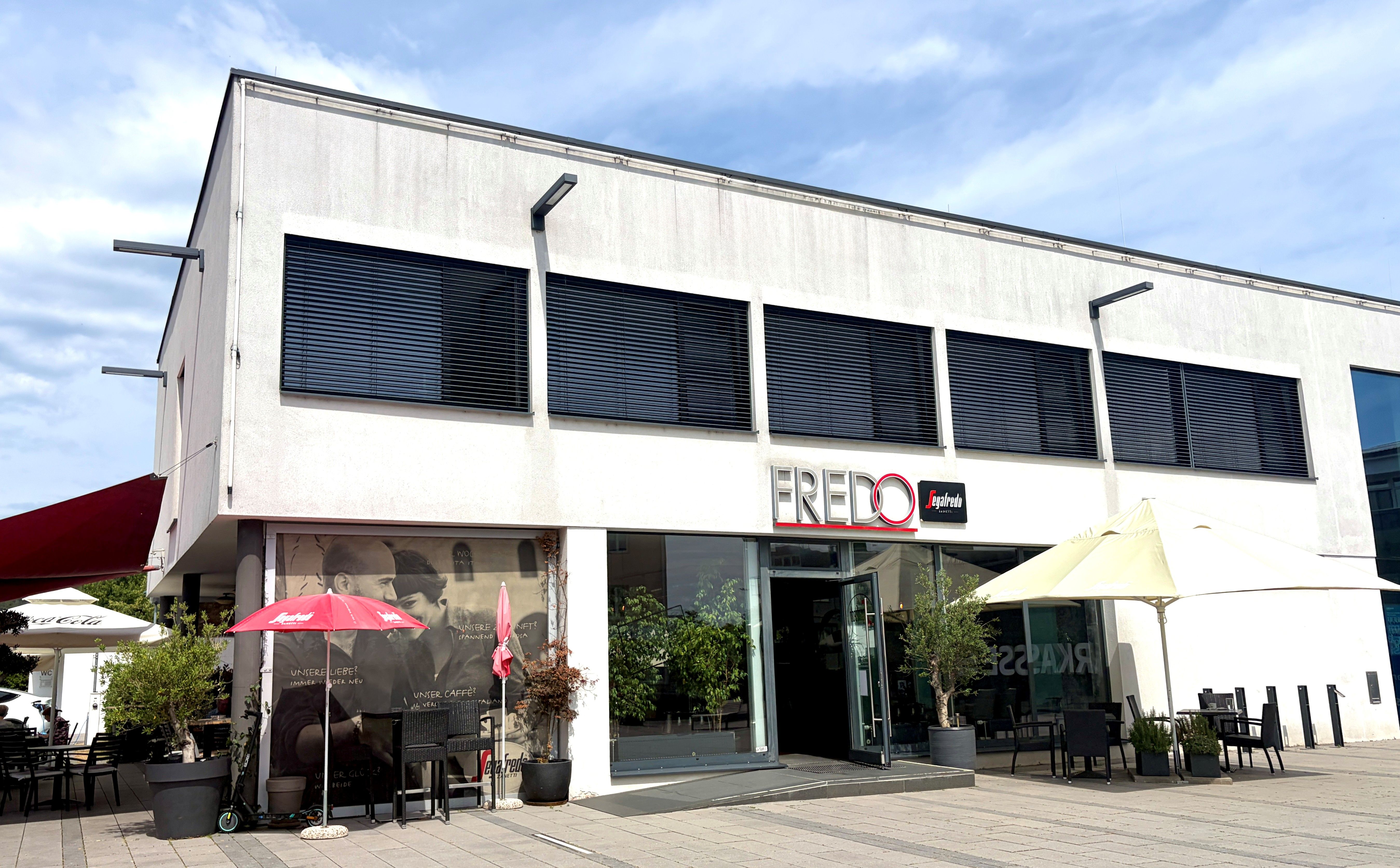 Außenansicht eines modernen Cafés mit dem Namen Fredo, ausgestattet mit Sonnenschirmen und Sitzgelegenheiten im Freien.