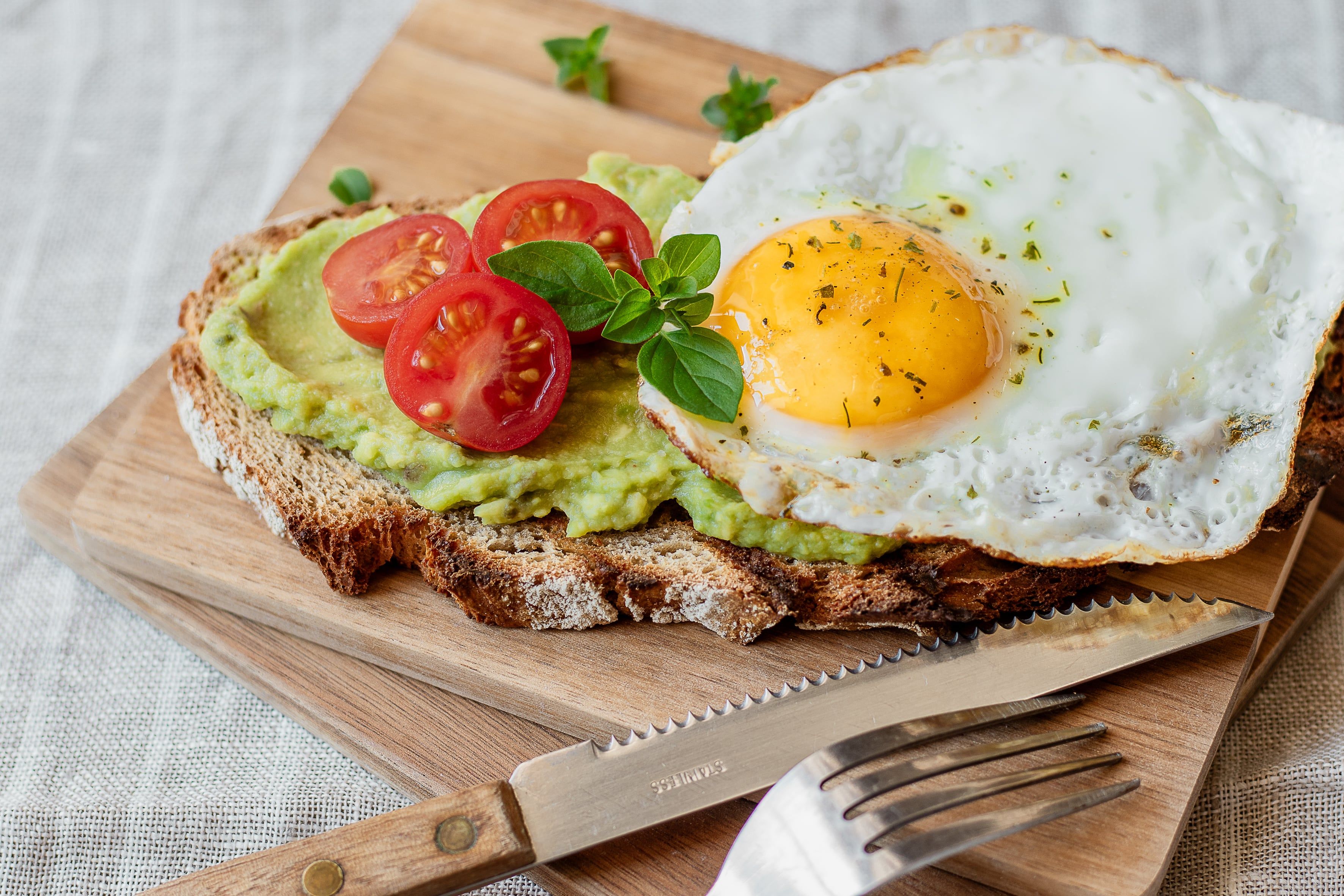 Ein belegtes Brot mit Avocado, Tomaten und Spiegelei auf einem Holzbrett.
