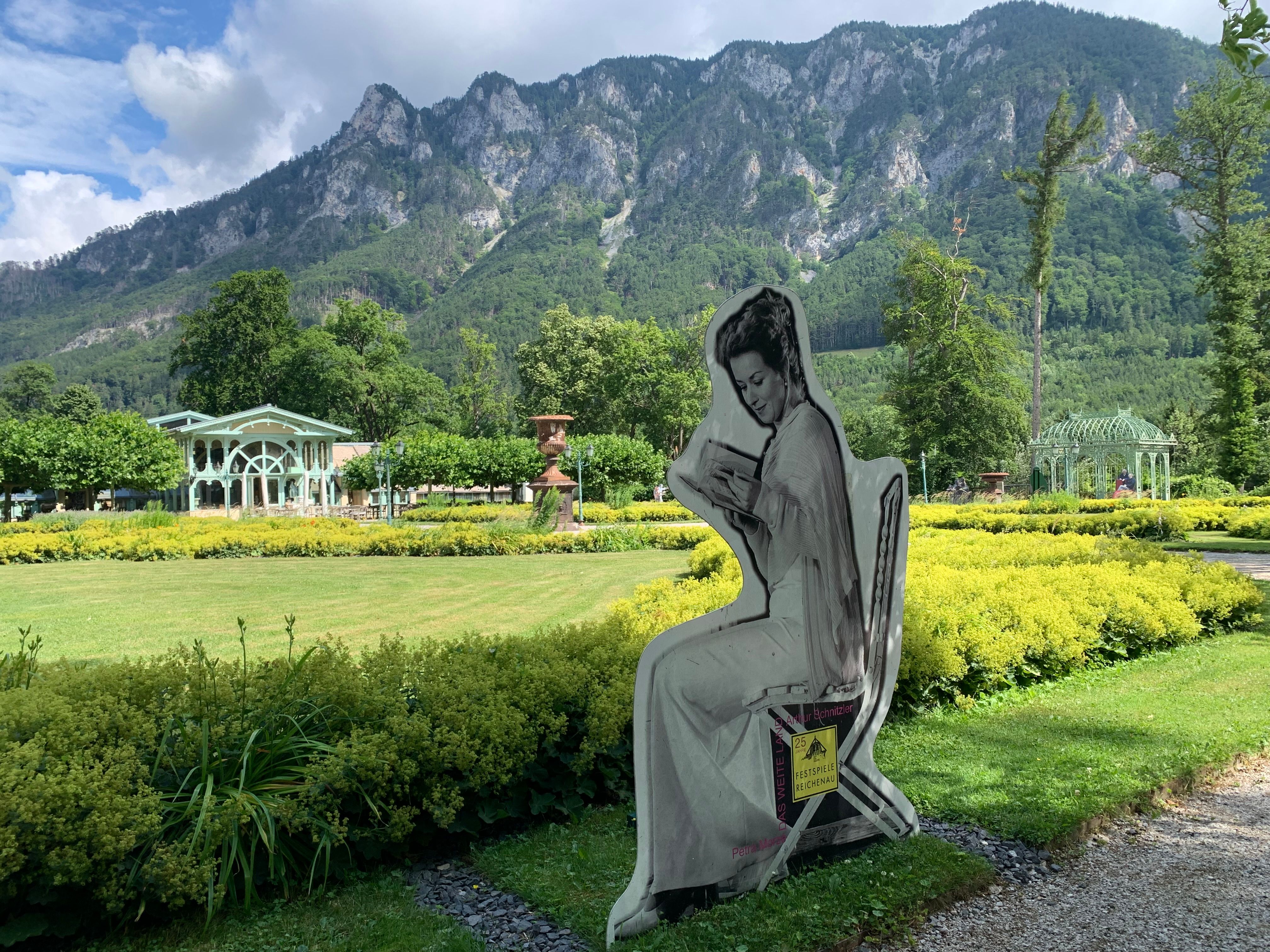 Gartenlandschaft mit Figur einer sitzenden Frau und Bergen im Hintergrund.