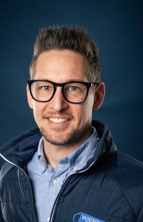 Portraitfoto Wiener Alpen Mitarbeiter, der eine Jacke mit Wiener Alpen Logo trägt und vor einem dunklem Hintergrund steht.