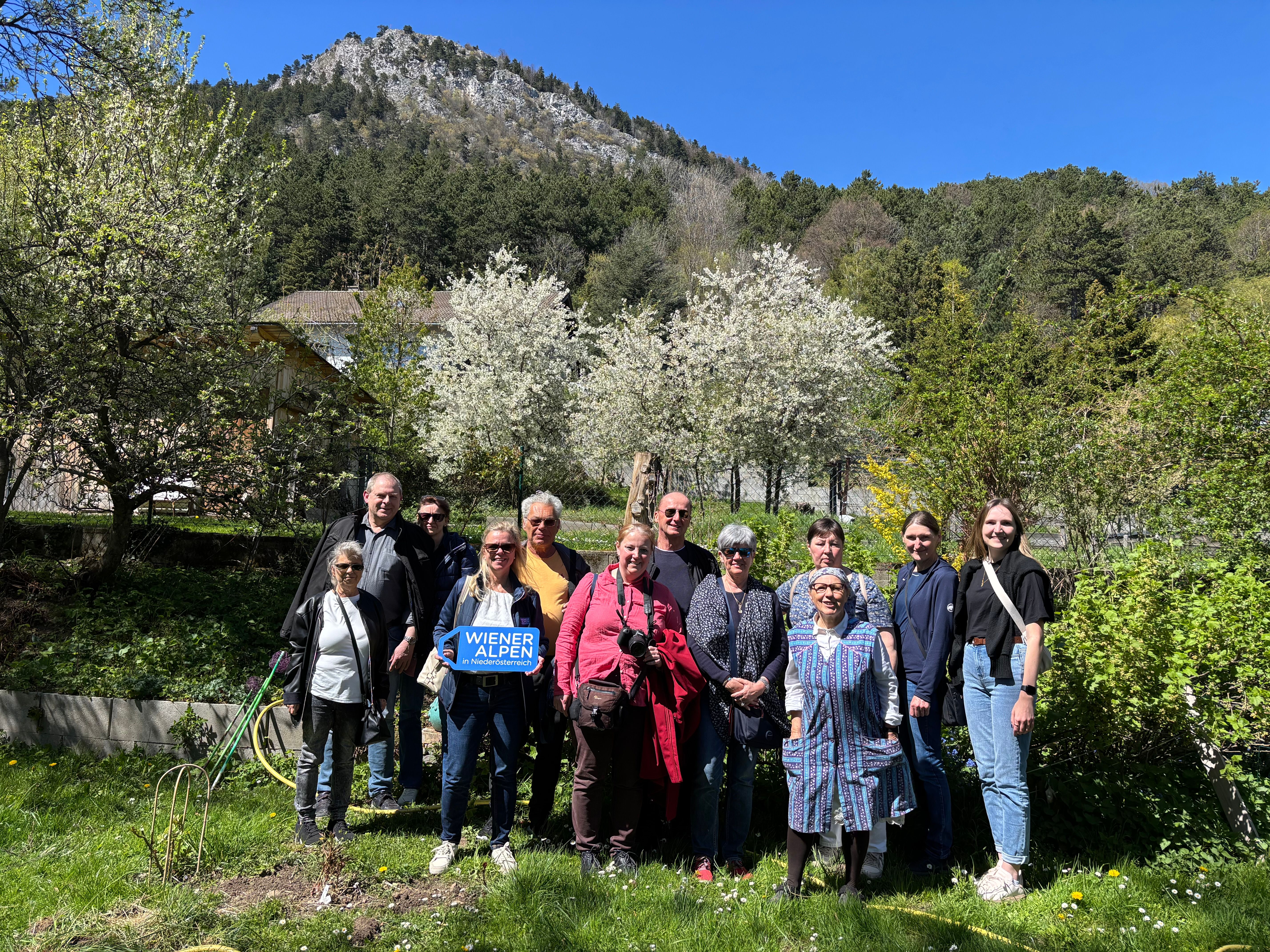Eine Gruppe von rund 12 Personen steht in der Wiese in einem grünen Garten bei Sonnenschein. Eine Frau hält ein Wiener Alpen Logo-Schild in der Hand.