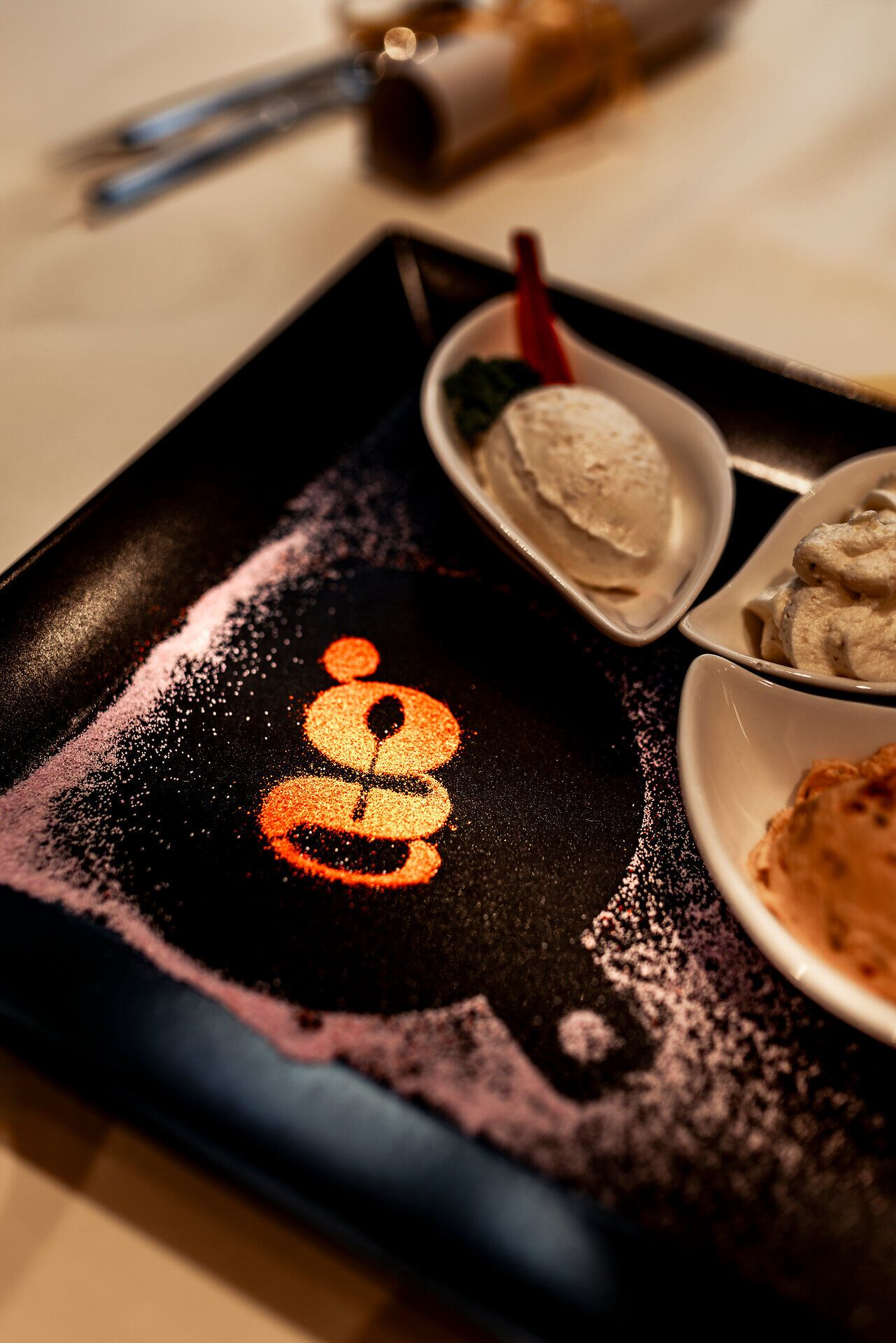 Dessertvariation mit Eiscreme, verziert mit Zucker und dem Buchstaben "g" auf einem Teller.