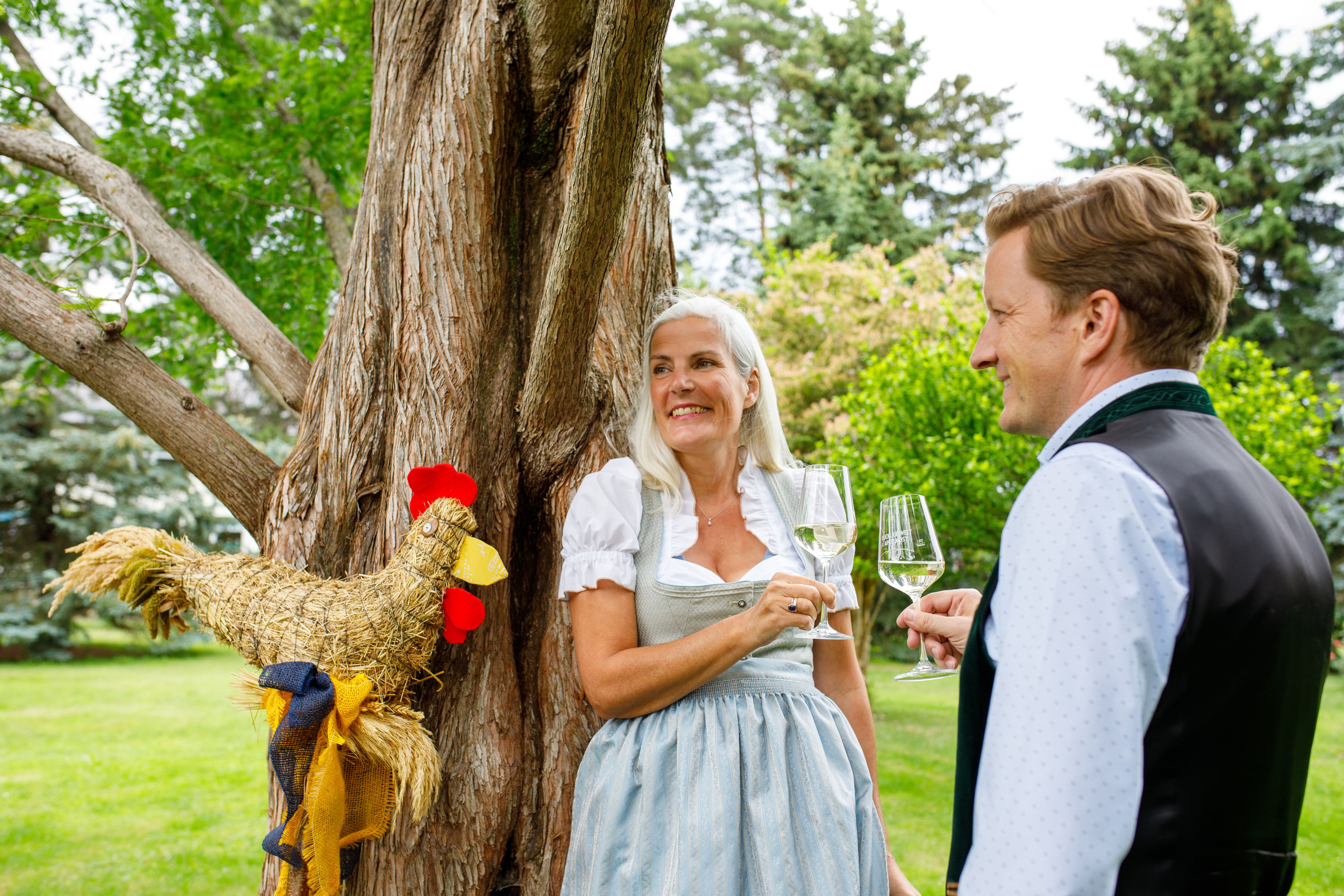 Eine Frau und ein Mann in Tracht stoßen mit Weingläsern an. Daneben ein Baum und ein Schnidahahn.