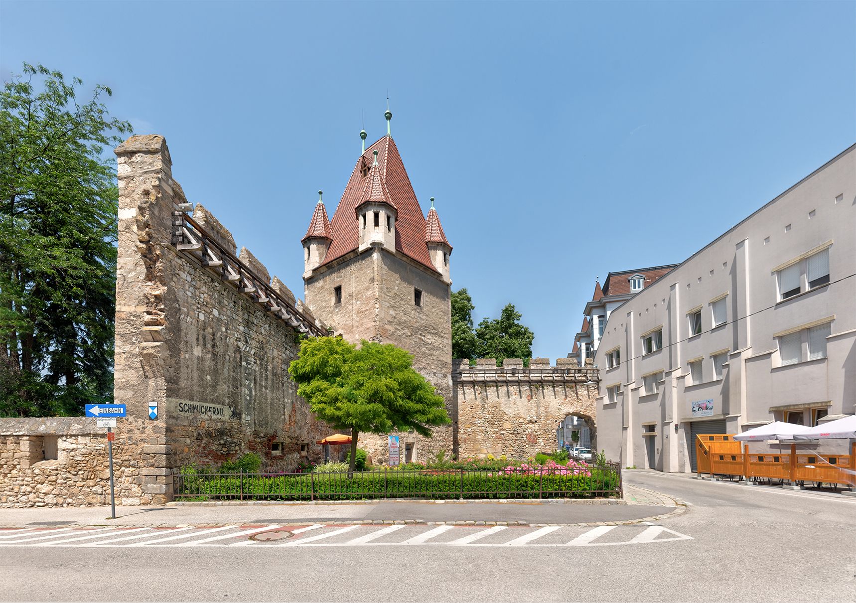Historischer Reckturm mit rotem Dach und angrenzender Mauer, umgeben von modernen Gebäuden und einer Straße.