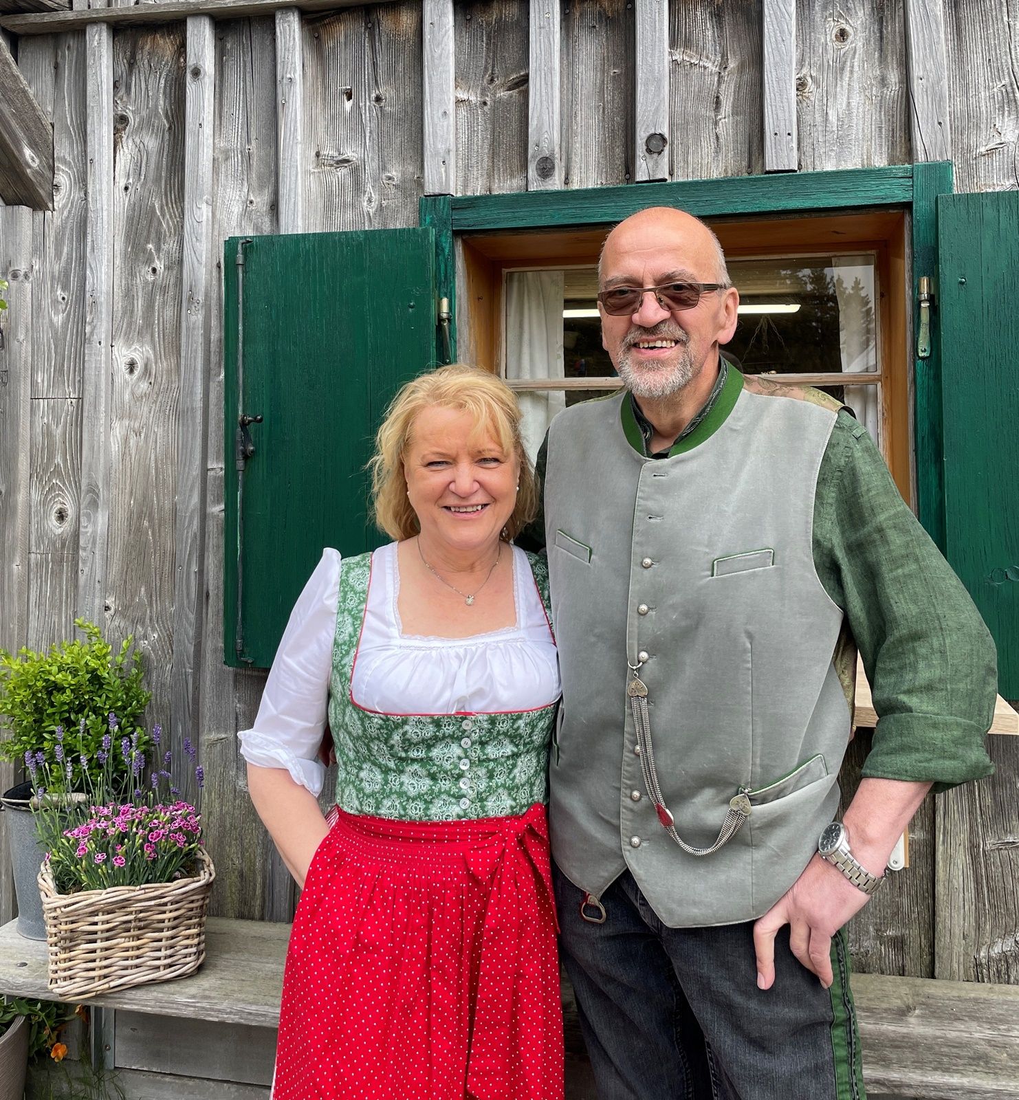 Ein Mann und eine Frau in traditioneller Tracht vor einem Holzhaus mit grünen Fensterläden.