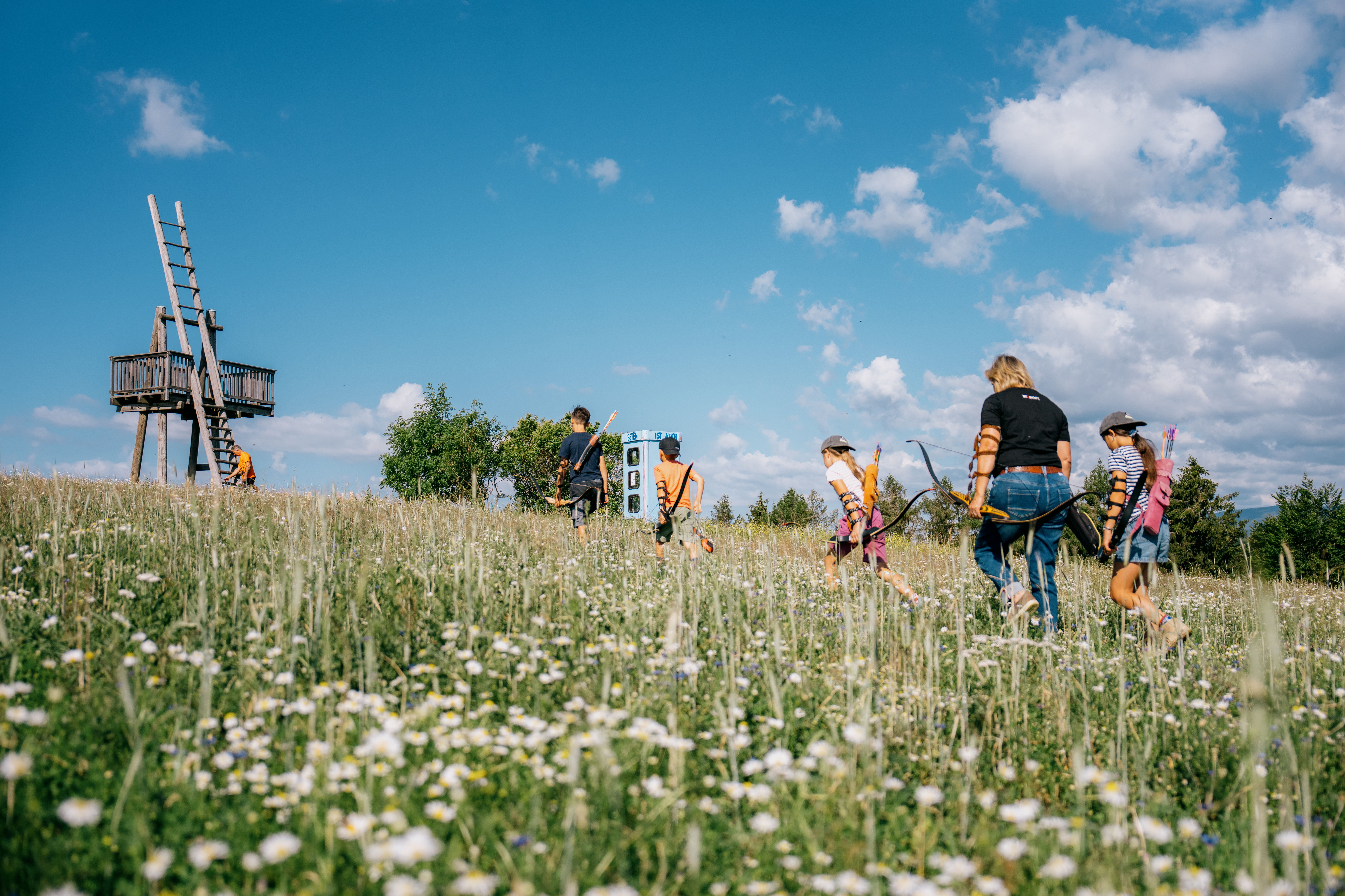 Eine Gruppe von Menschen mit Pfeilen und Bögen geht über eine blühende Wiese in Richtung eines hölzernen Aussichtsturms unter blauem Himmel.