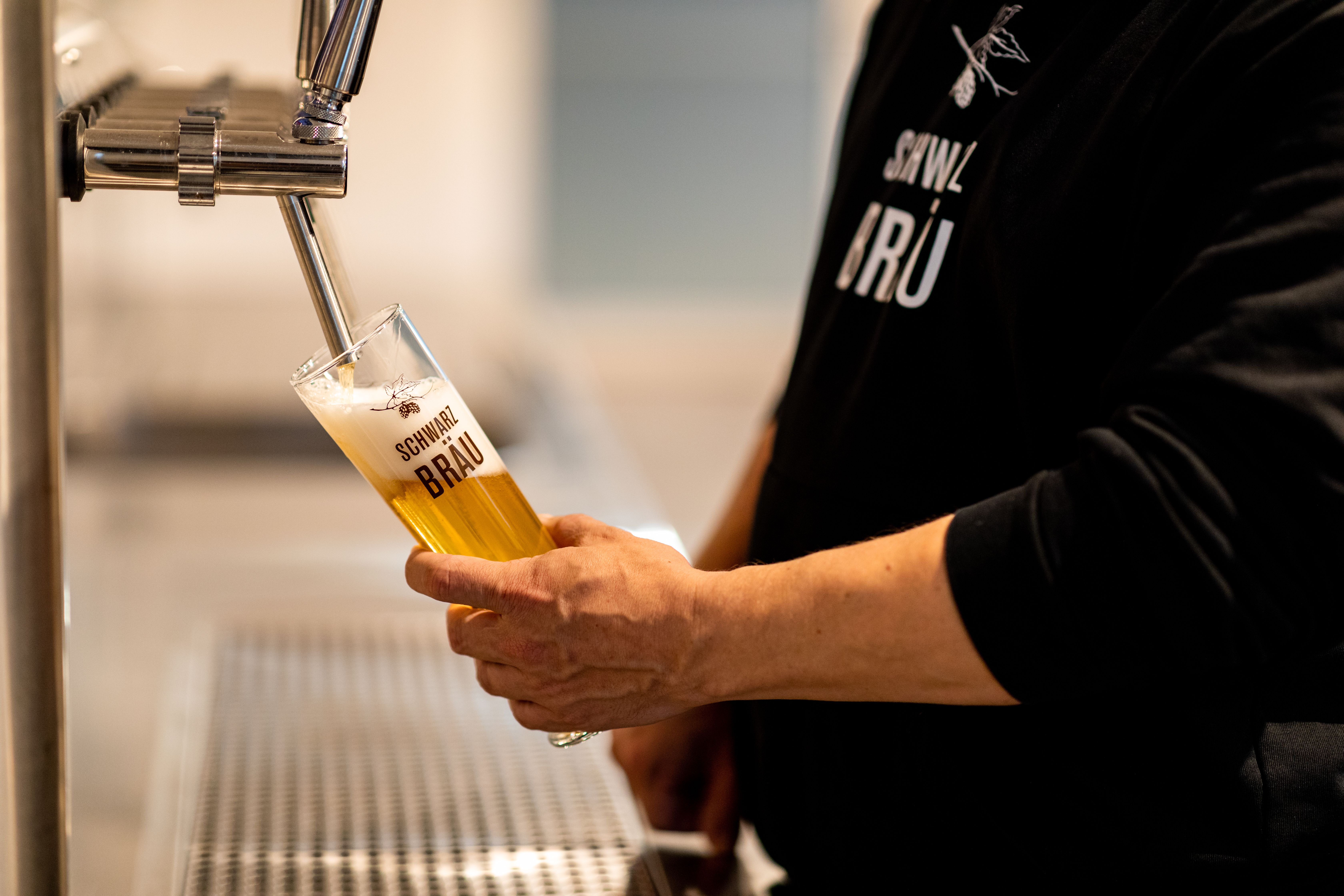 Jemand zapft Bier von einer Zapfanlage aus Edelstahl. Das Glas ist hoch und schmal, das Logo der Brauerei Schwarzbräu ist in dunkelem Braun auf das Glas gedruckt.