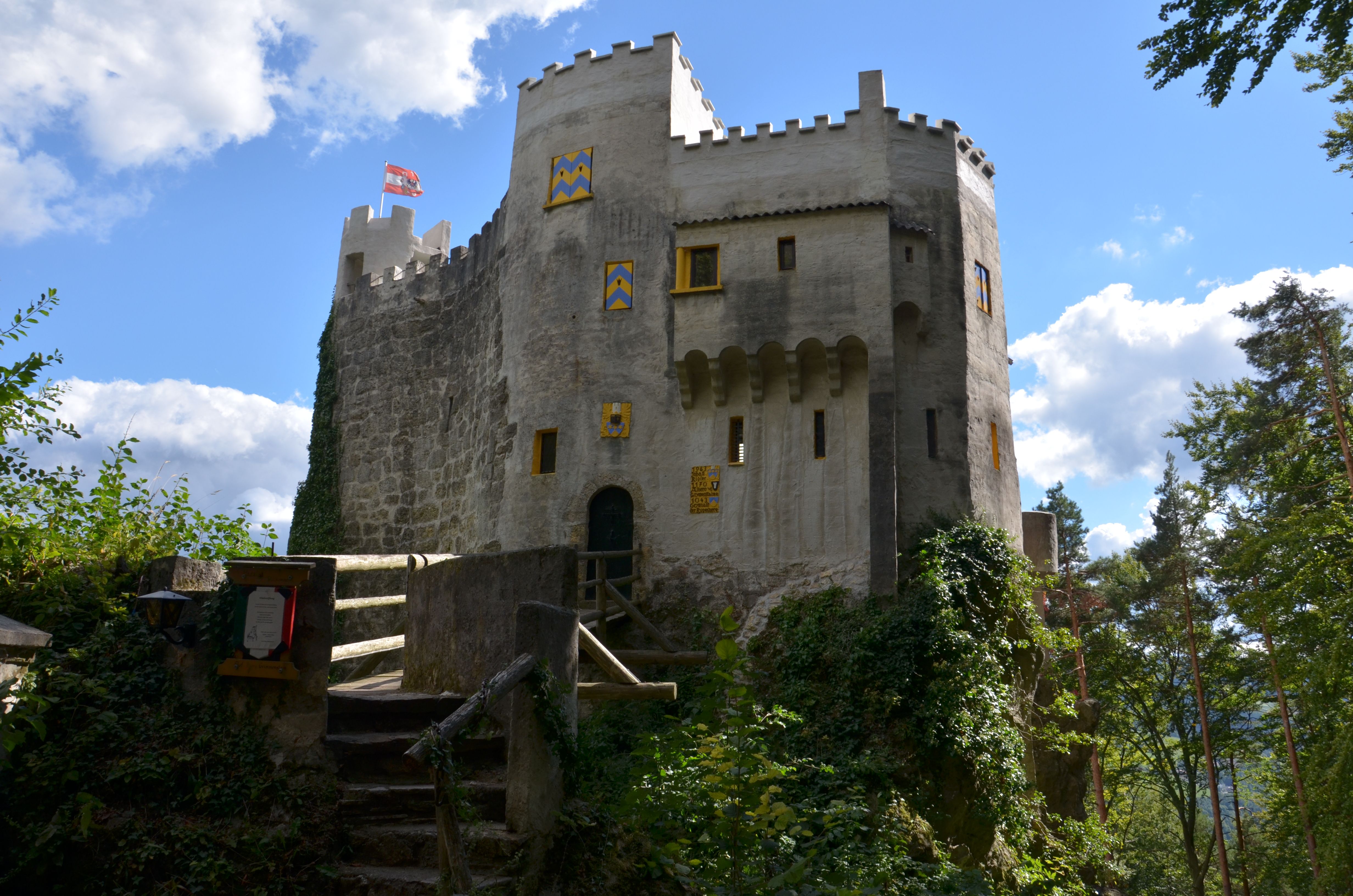 Burg Grimmenstein auf einem Hügel, mit Zinnen, Fahne und Wald im Vordergrund unter blauem Himmel. Stufen und eine Brücke führen zum Eingangstor.
