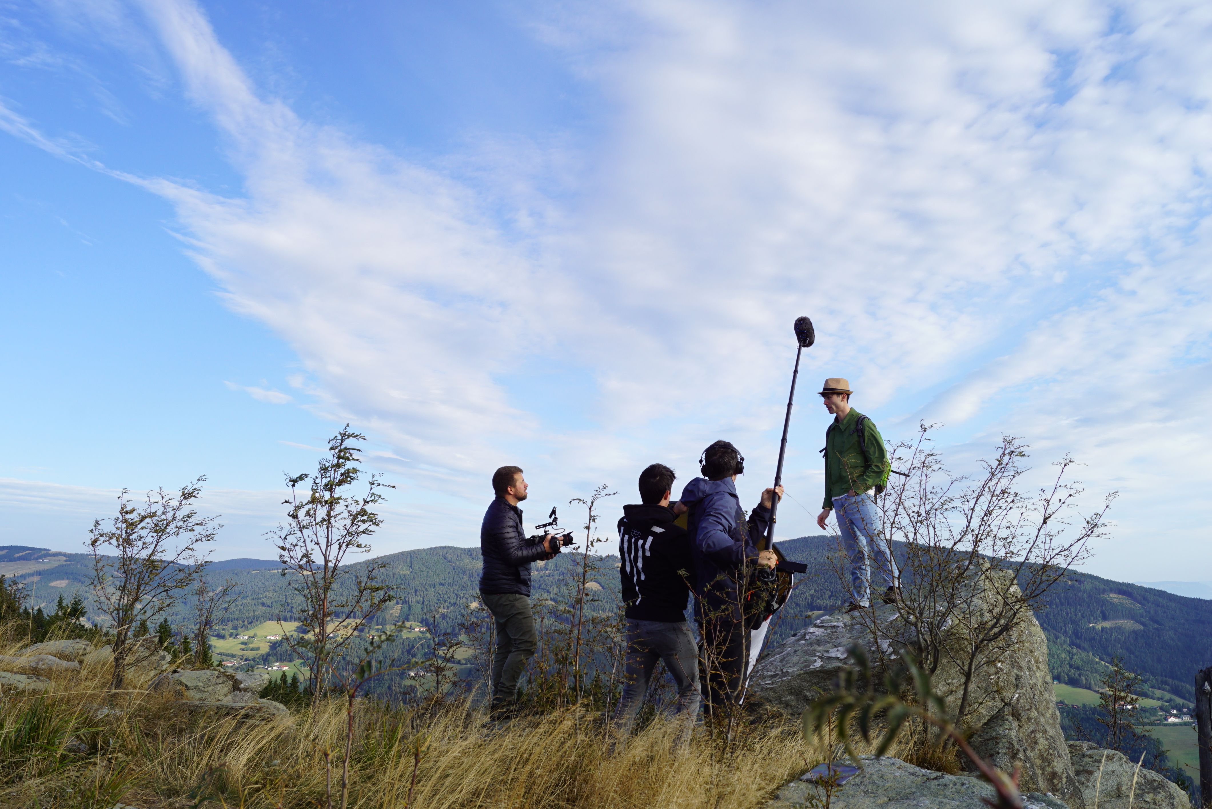 Bei Filmaufnahmen steht ein Darsteller vor hügeliger Landschaft, vor ihm stehen Kameramann, Tonmann mit langem Mikrofon und ein weiterer Mann.