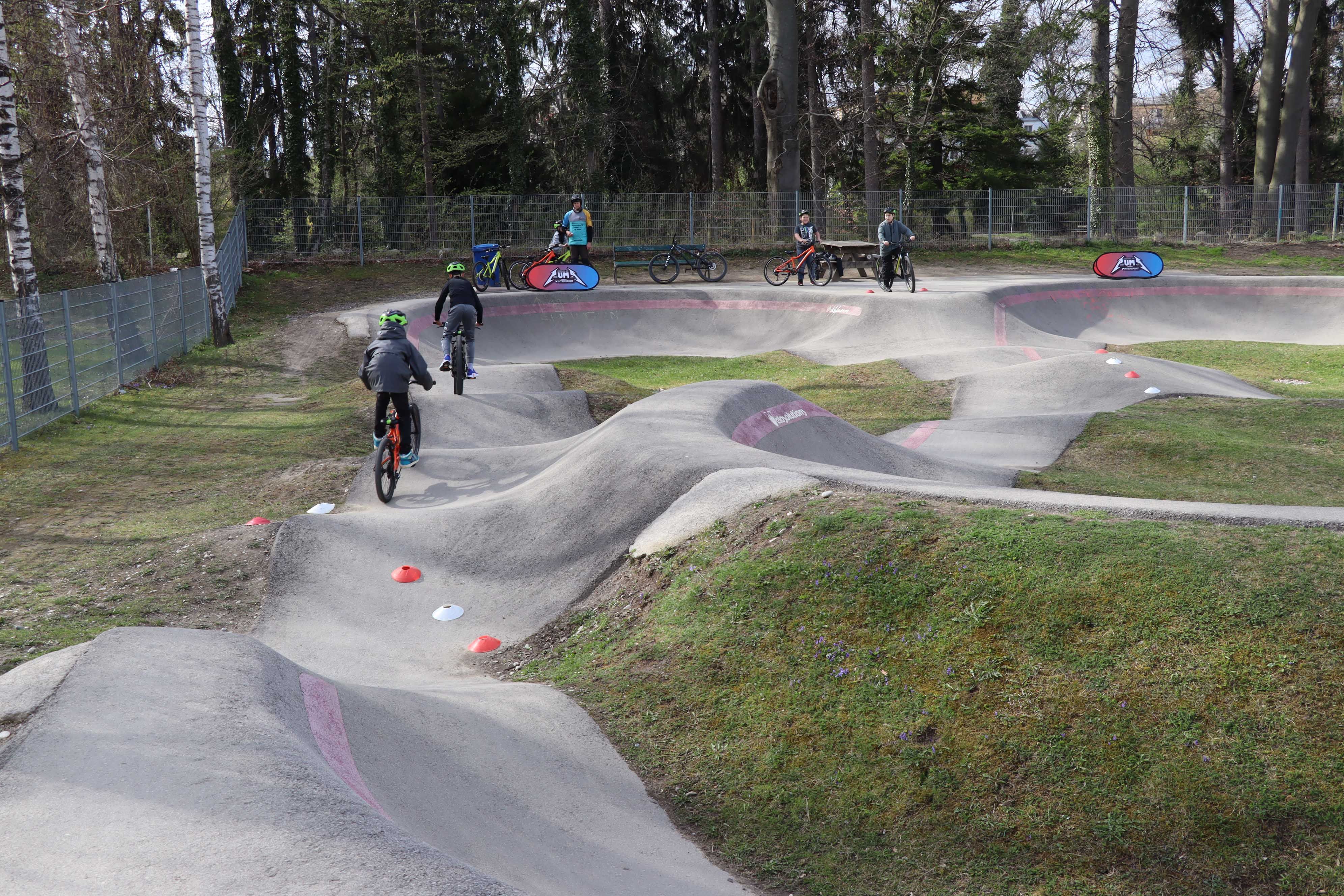 Kinder und Jugendliche fahren mit Mountainbikes auf einem Pumptrack mit Wellen und Kurven, während weitere Personen mit Fahrrädern am Rand stehen und zusehen. Die Anlage liegt in einer grünen Parklandschaft mit Bäumen.