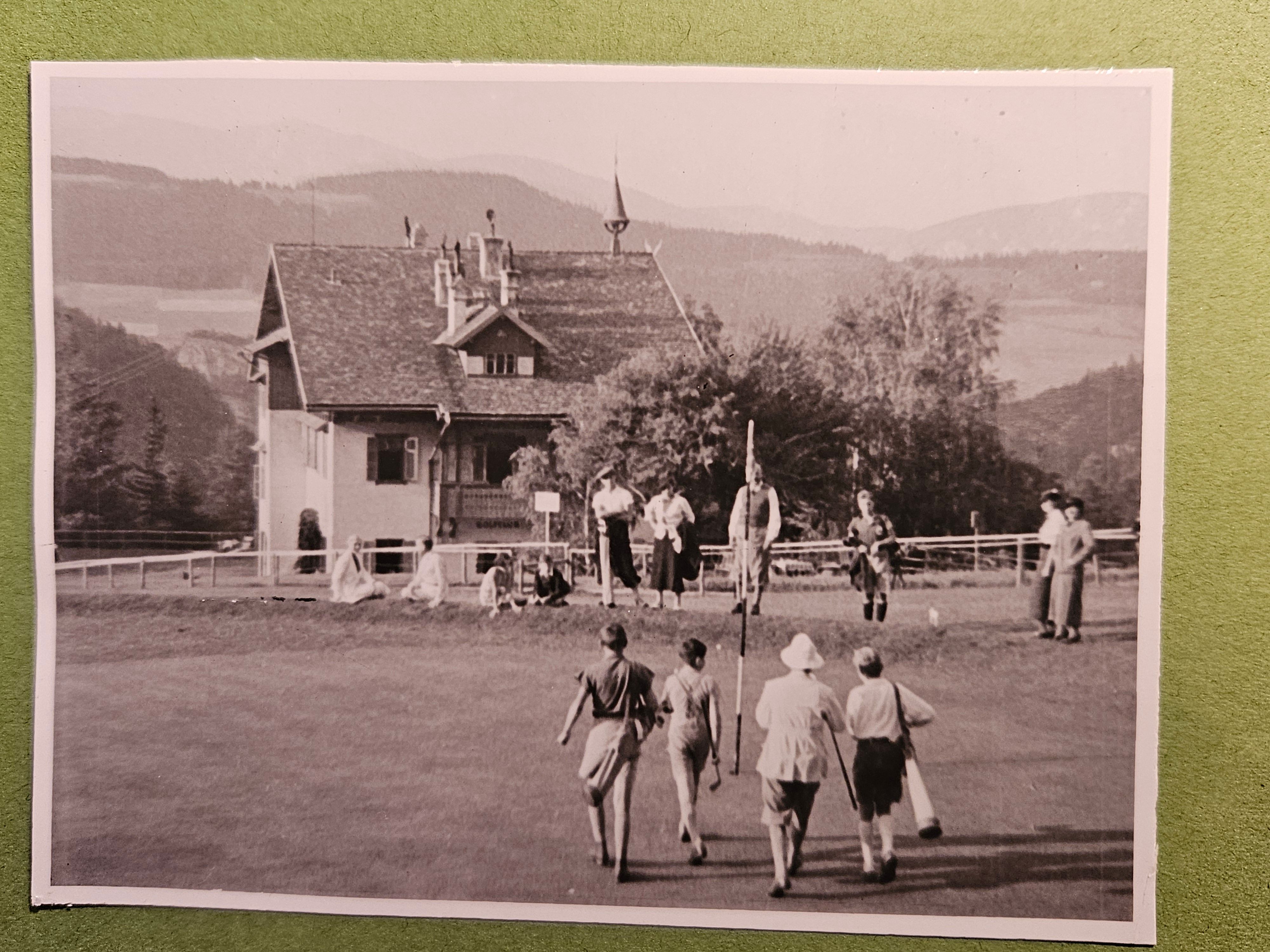 Historisches Schwarz-Weiß-Foto des Golfplatzes am Semmering mit Golfern auf dem Grün und traditionellem Gebäude im Hintergrund, eingebettet in die alpine Landschaft.