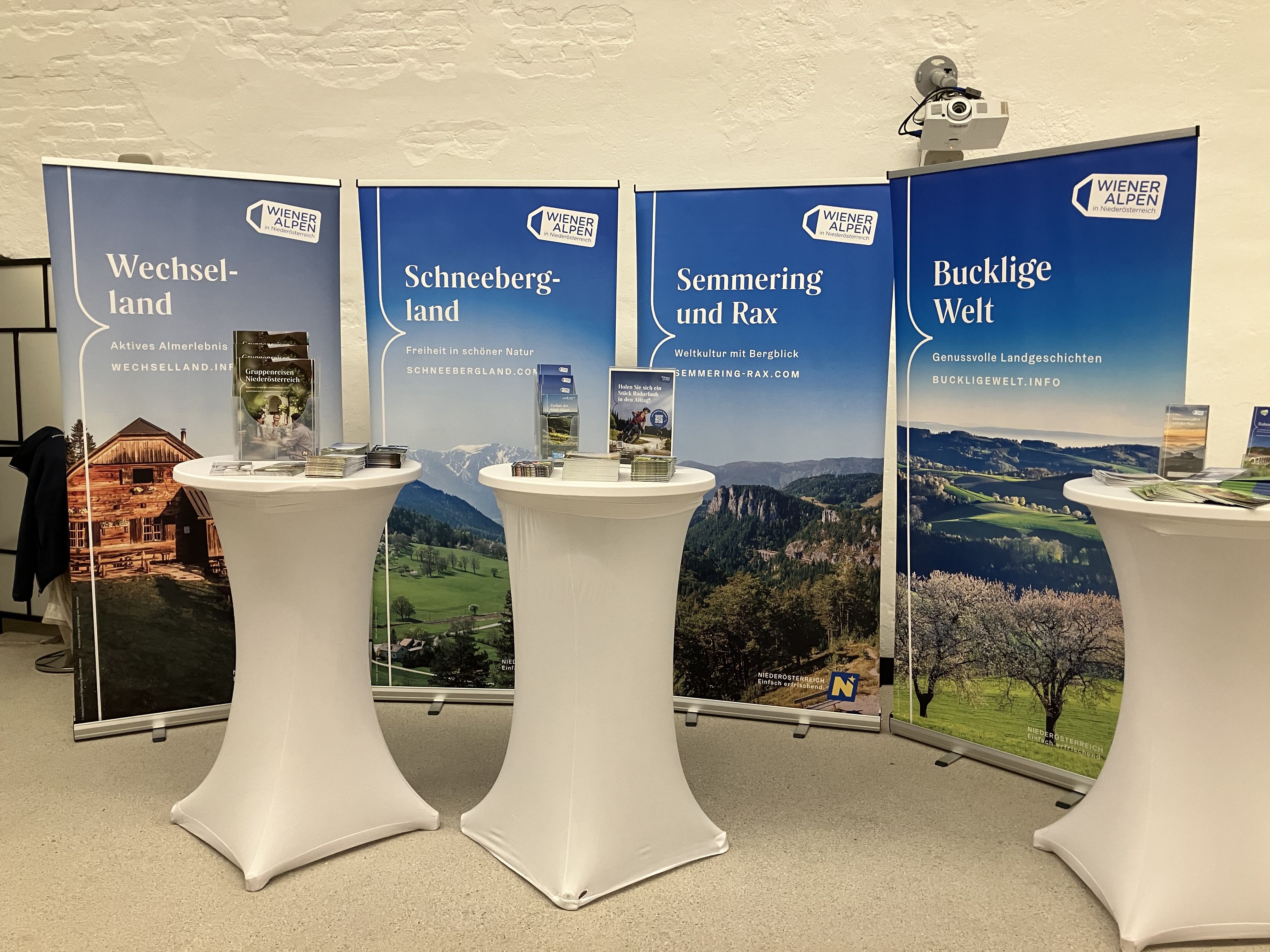 Vor vier Rückwänden einzelner Regionen der Wiener Alpen stehen drei Stehtische, auf denen verschiedene Broschüren angeboten werden. 